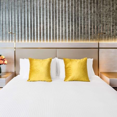 Radisson Blu Plaza Hotel ซิดนีย์ ภายนอก รูปภาพ The photo shows a stylish hotel room with a modern design. In the center, there's a neatly made bed with a white bedspread and two decorative yellow pillows. The headboard has a sleek, contemporary look, and there are two bedside tables on either sid
