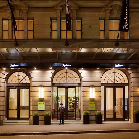 Radisson Blu Plaza Hotel ซิดนีย์ ภายนอก รูปภาพ The photo depicts the entrance of a Radisson hotel. The building features elegant architecture, with large arched windows and a well-lit canopy above the entrance. There are two potted plants on either side of the doors, which are framed by modern li
