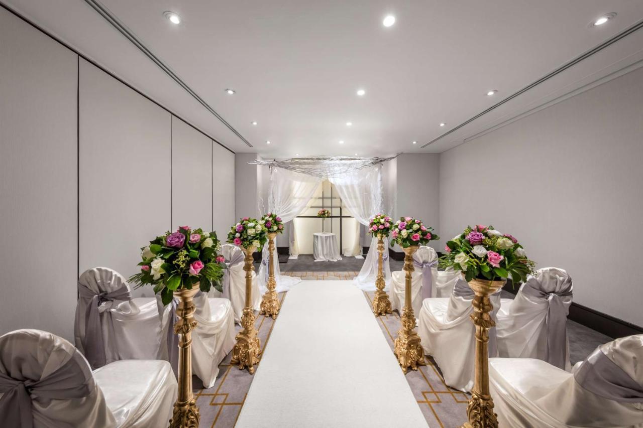 Radisson Blu Plaza Hotel ซิดนีย์ ภายนอก รูปภาพ The photo shows a beautifully arranged indoor event space, likely set up for a wedding ceremony. There is a long white aisle runner leading to a decorated archway at the end. On either side of the aisle, there are elegant chairs draped in white fabri