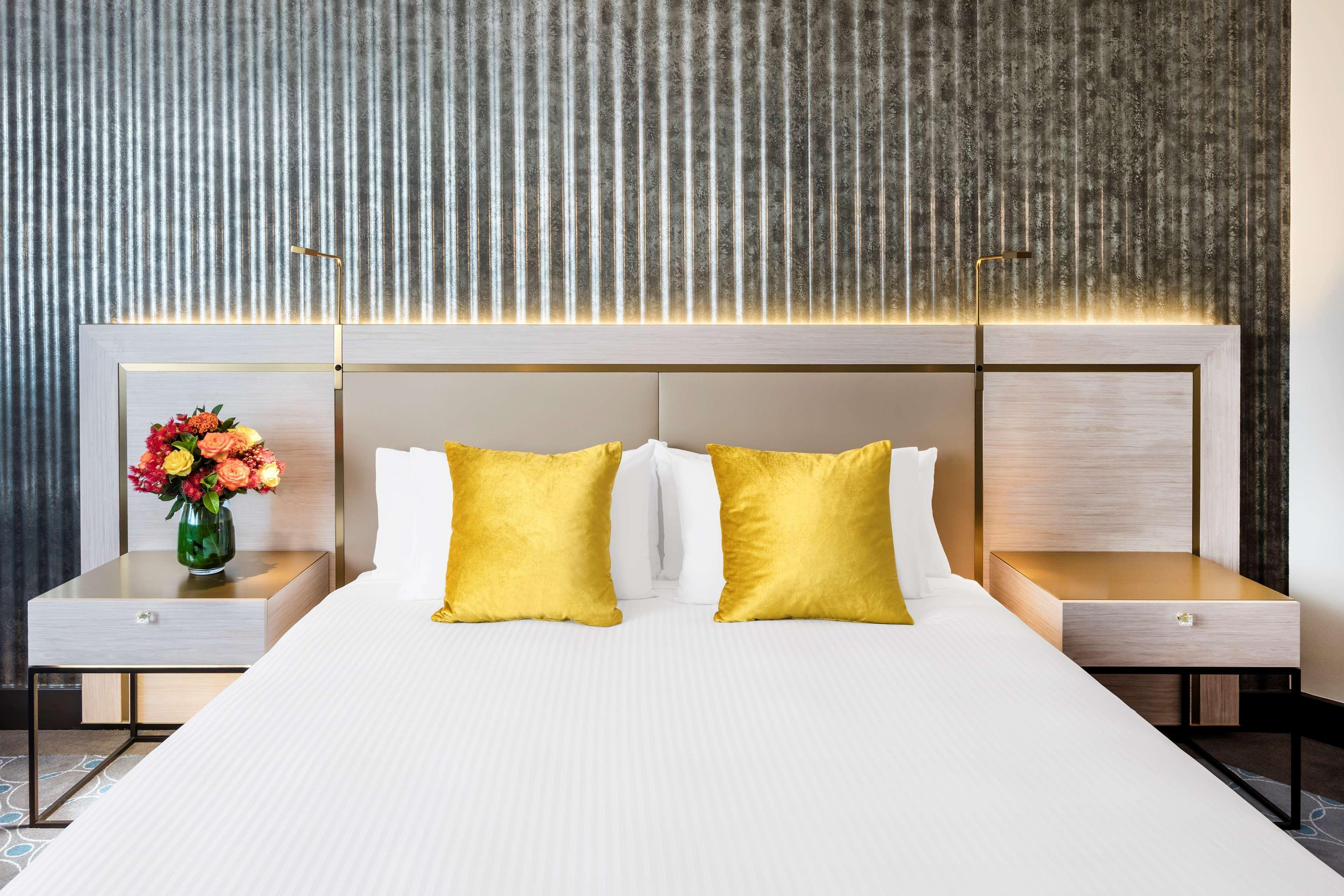 Radisson Blu Plaza Hotel ซิดนีย์ ภายนอก รูปภาพ The photo shows a stylish hotel room with a modern design. In the center, there's a neatly made bed with a white bedspread and two decorative yellow pillows. The headboard has a sleek, contemporary look, and there are two bedside tables on either sid