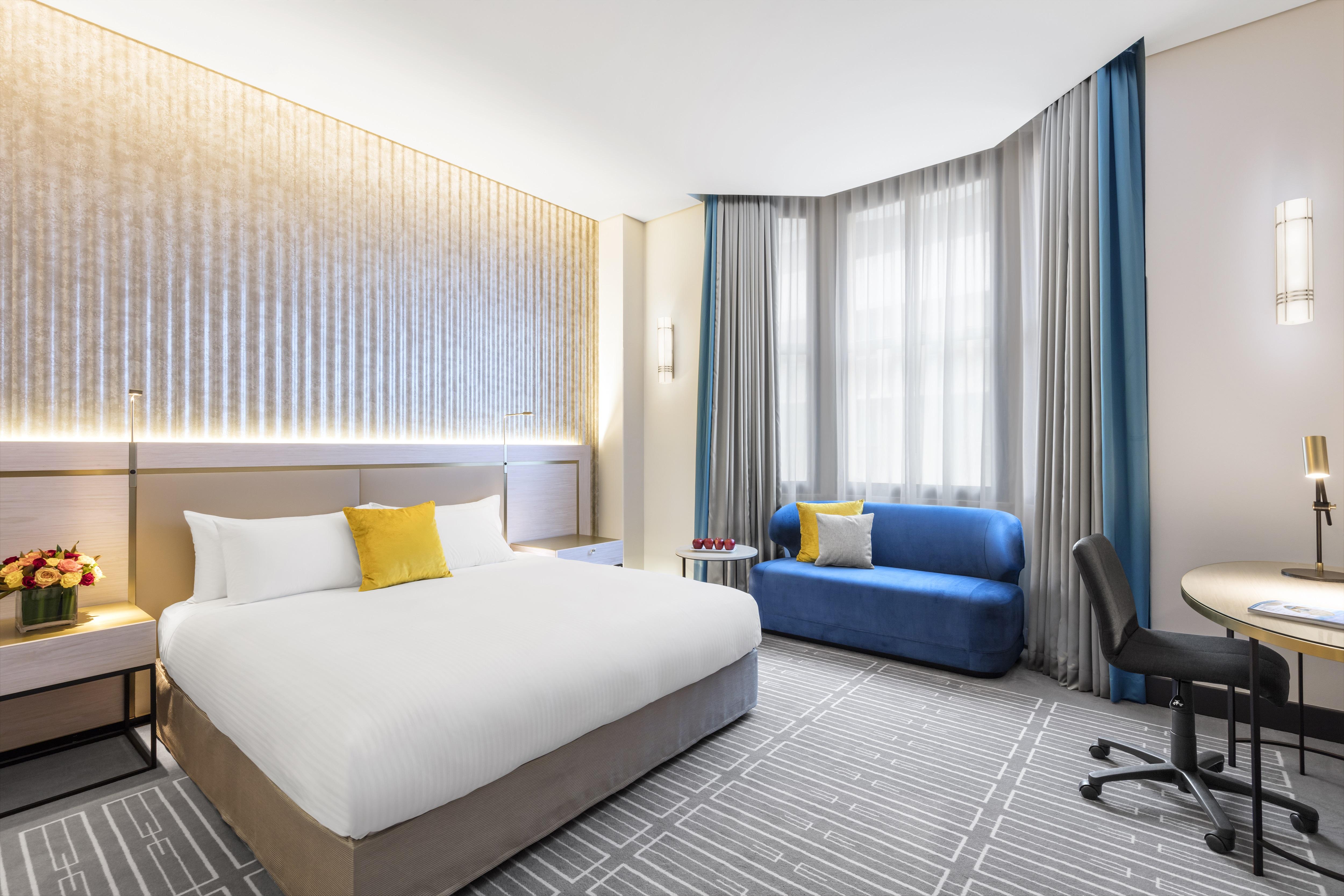 Radisson Blu Plaza Hotel ซิดนีย์ ภายนอก รูปภาพ The photo shows a modern hotel room featuring a large bed with white linens and a yellow accent pillow. To the side, there is a blue sofa and a small round table with a chair positioned next to it. The room is well-lit, with natural light coming thro