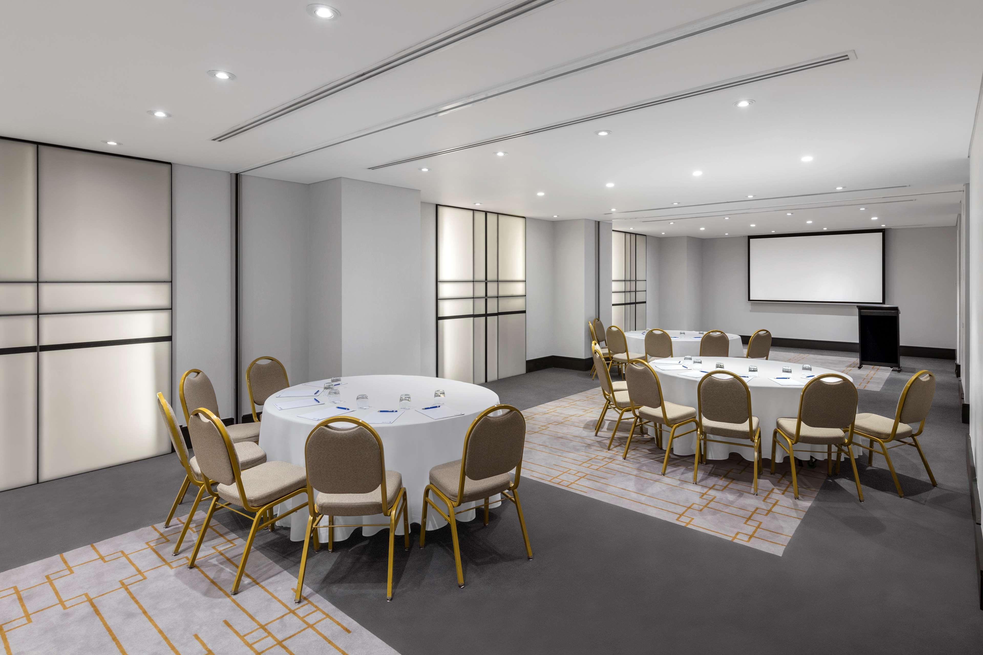 Radisson Blu Plaza Hotel ซิดนีย์ ภายนอก รูปภาพ The photo shows a modern meeting room or conference space. It features several round tables covered with white tablecloths, surrounded by gold-colored chairs. The walls are light-colored, and there are large, sliding partitions that could separate th