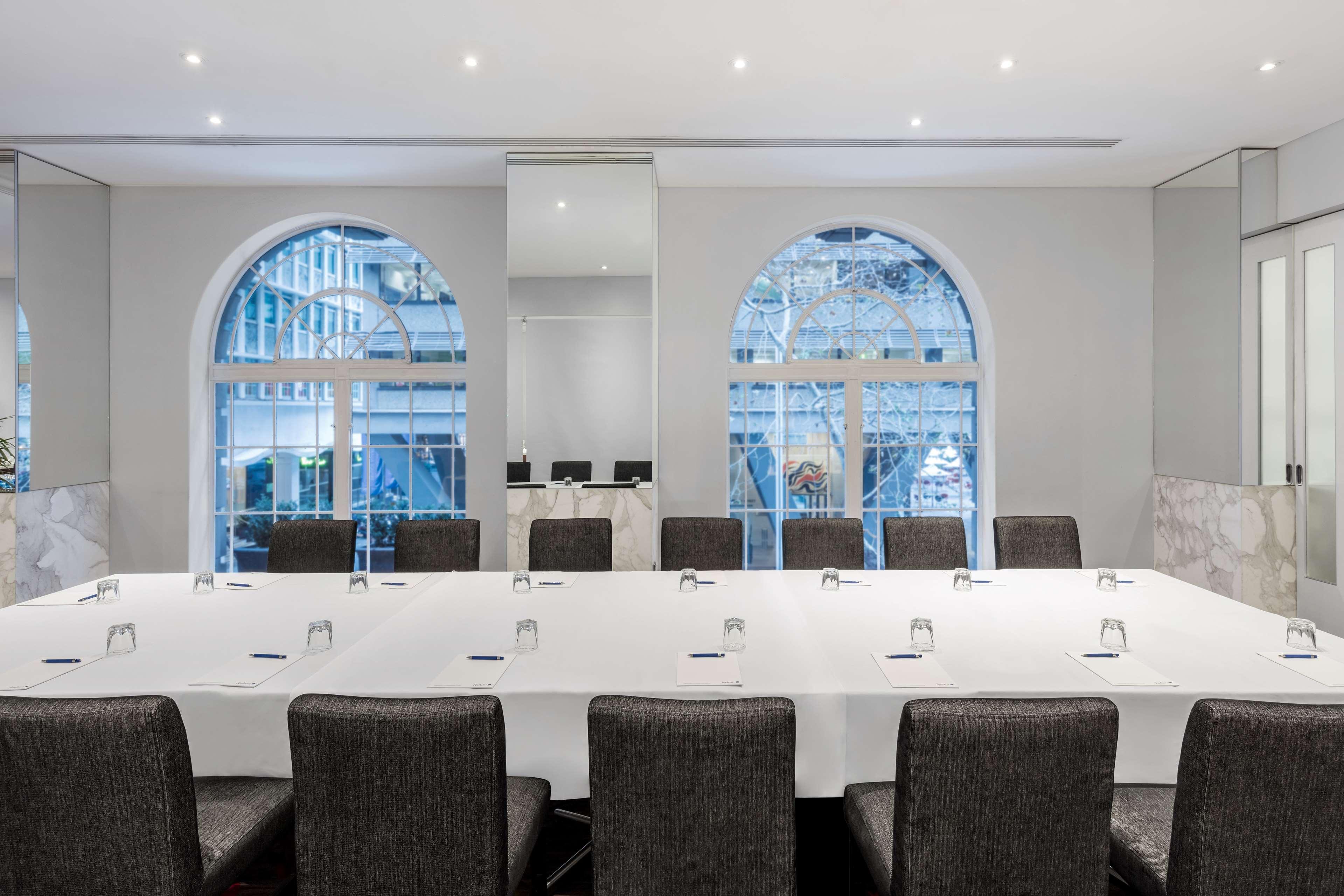 Radisson Blu Plaza Hotel ซิดนีย์ ภายนอก รูปภาพ The image shows a conference room set up for a meeting. It features a long table covered with a white tablecloth, arranged with several place settings and water glasses. Around the table are black, padded chairs. The room has large arched windows tha
