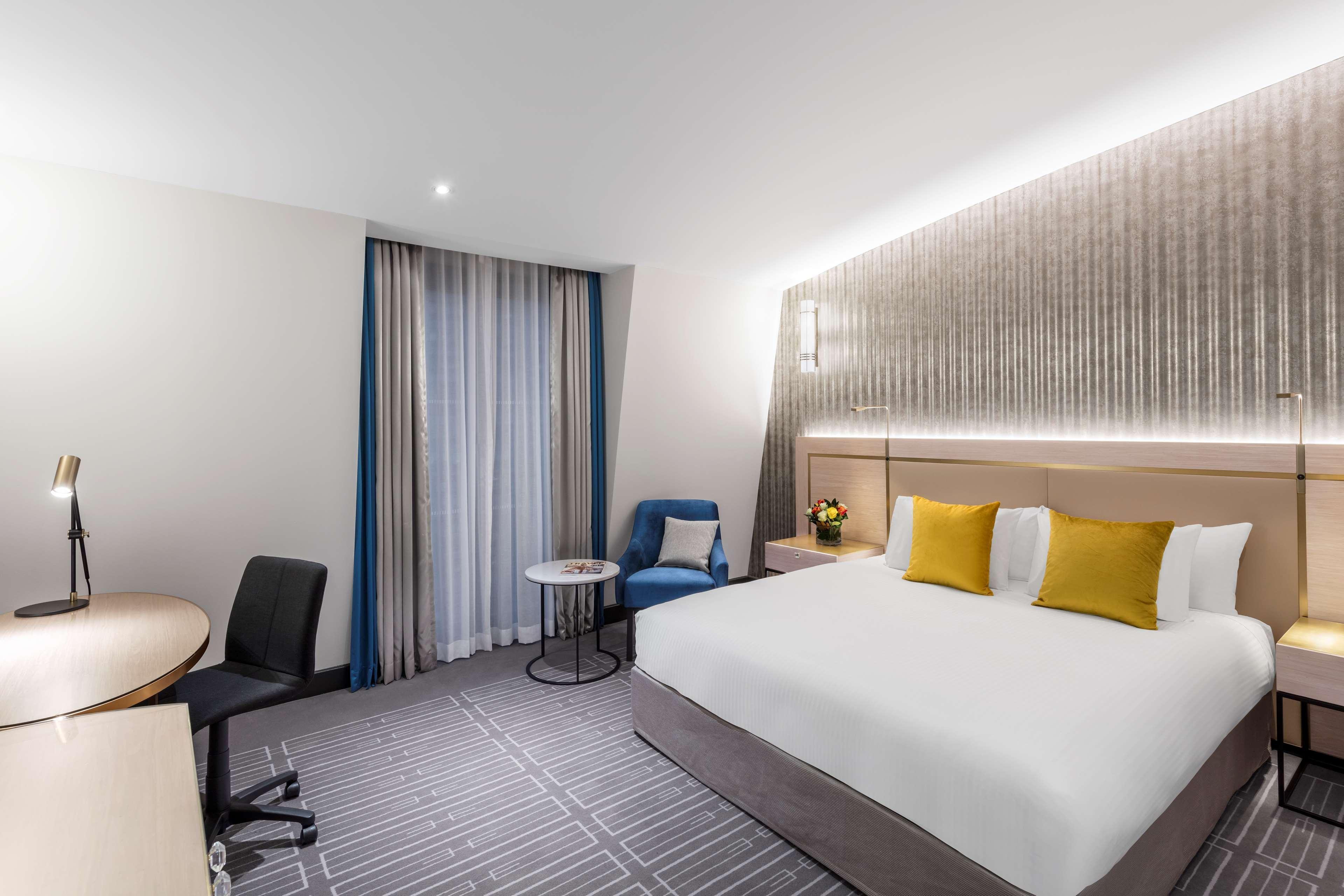Radisson Blu Plaza Hotel ซิดนีย์ ภายนอก รูปภาพ The photo shows a modern hotel room featuring a large bed with white linens and yellow accent pillows. There's a plush blue chair next to a small round table. To one side, a desk with a black office chair is positioned, likely for work or study. The 