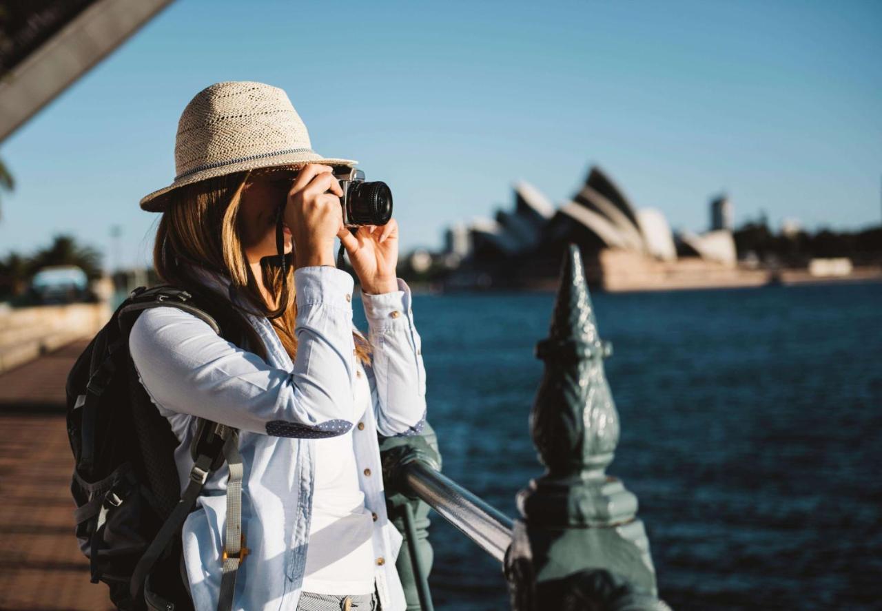 Radisson Blu Plaza Hotel ซิดนีย์ ภายนอก รูปภาพ The photo shows a woman standing by a railing near a body of water, taking a picture with a camera. She is wearing a light-colored hat and a backpack, dressed in a casual outfit. In the background, the iconic Sydney Opera House is visible, along with