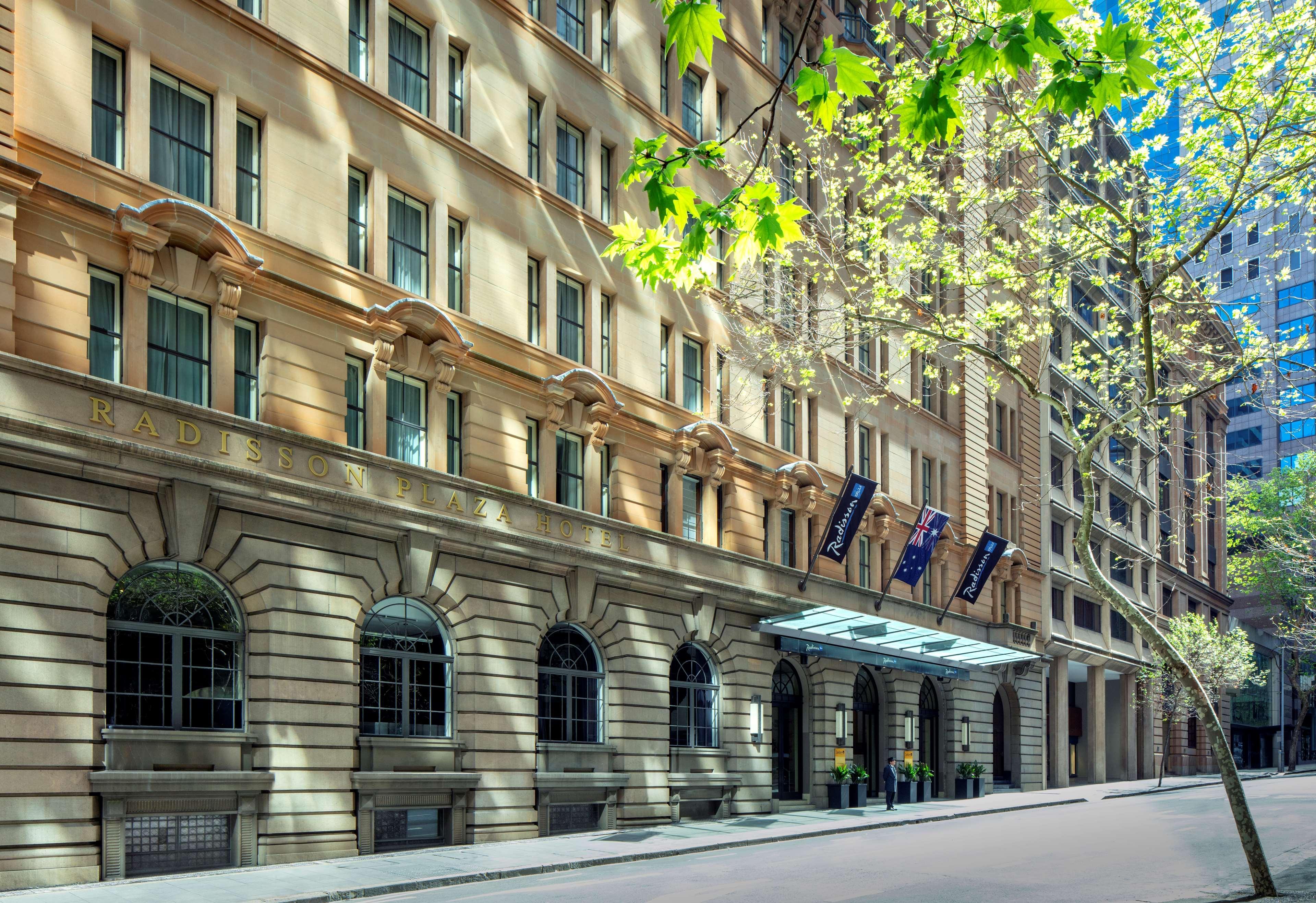 Radisson Blu Plaza Hotel ซิดนีย์ ภายนอก รูปภาพ The photo shows a well-maintained historic building with a classic architectural style. The façade features large arched windows and detailed stonework. The entrance is marked by a portico with a canopy, and there are banners hanging above. Trees are