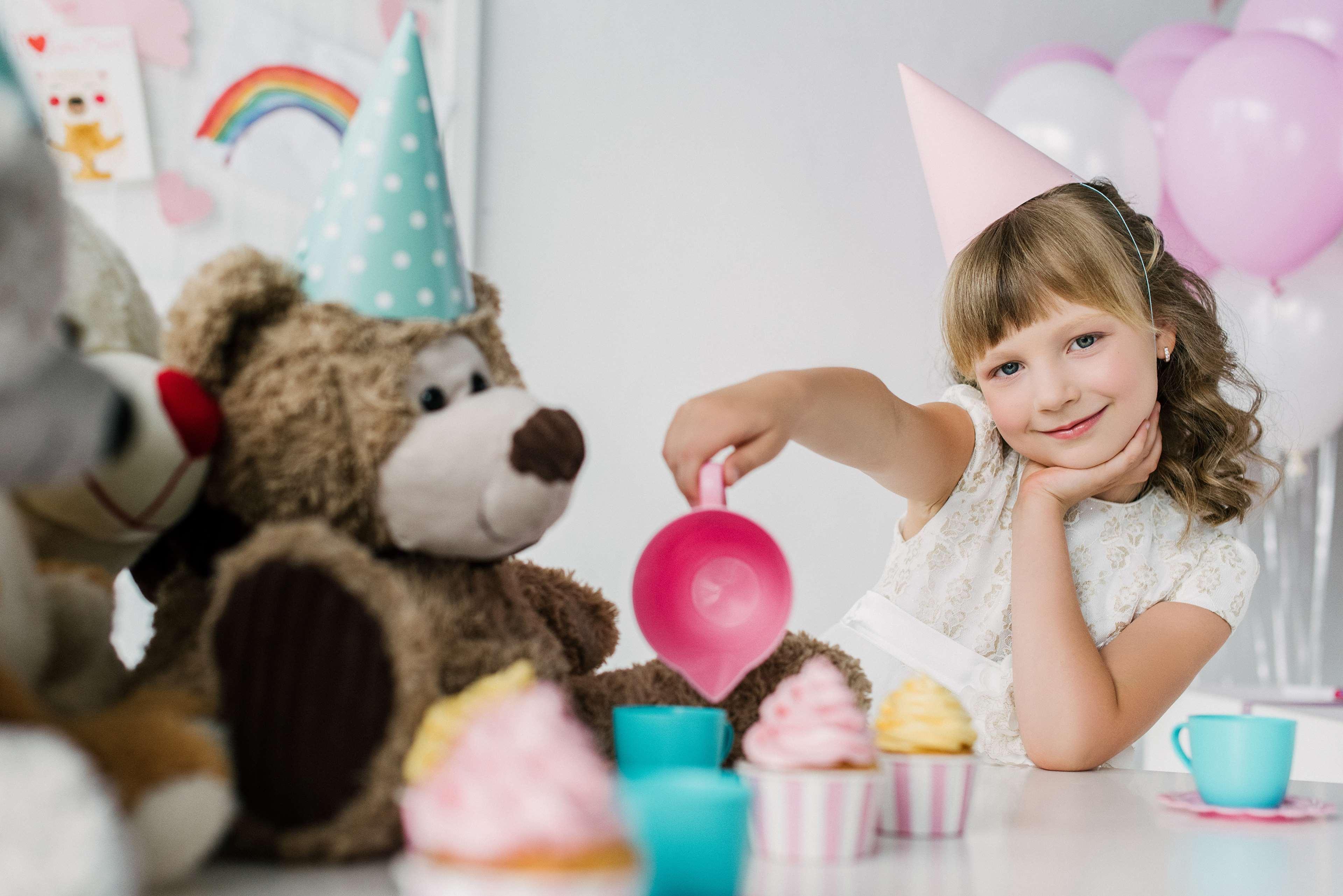 Radisson Blu Plaza Hotel ซิดนีย์ ภายนอก รูปภาพ The photo shows a young girl wearing a white dress and a pink party hat, sitting at a table. She is pouring something from a pink measuring cup into small cups. Beside her are two teddy bears, one wearing a brown color and the other with a white face