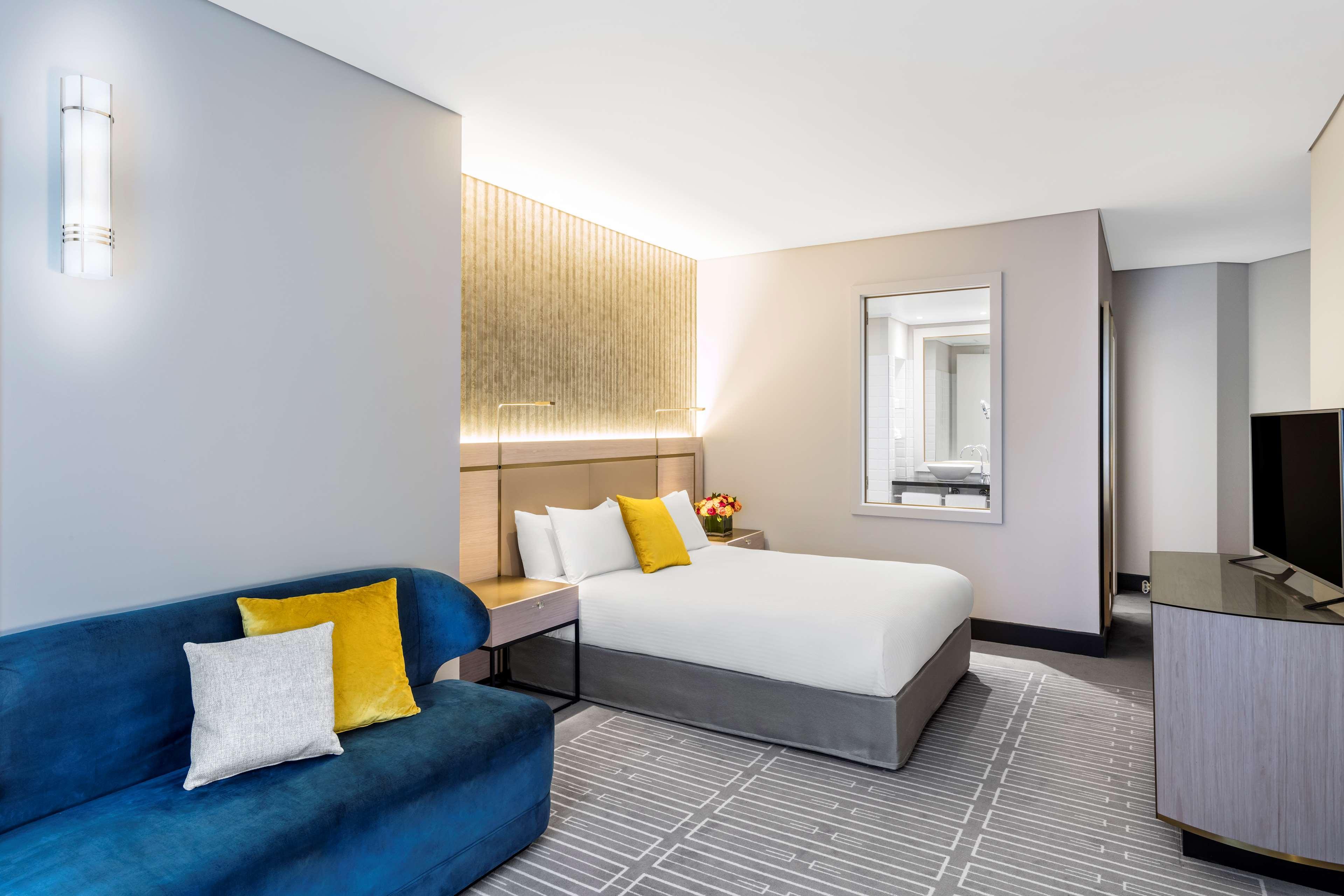 Radisson Blu Plaza Hotel ซิดนีย์ ภายนอก รูปภาพ The photo shows a modern hotel room. It features a double bed with white bedding and a grey upholstered base. To the left, there is a blue sofa accompanied by decorative pillows in yellow and white. A small wooden side table sits beside the sofa. The