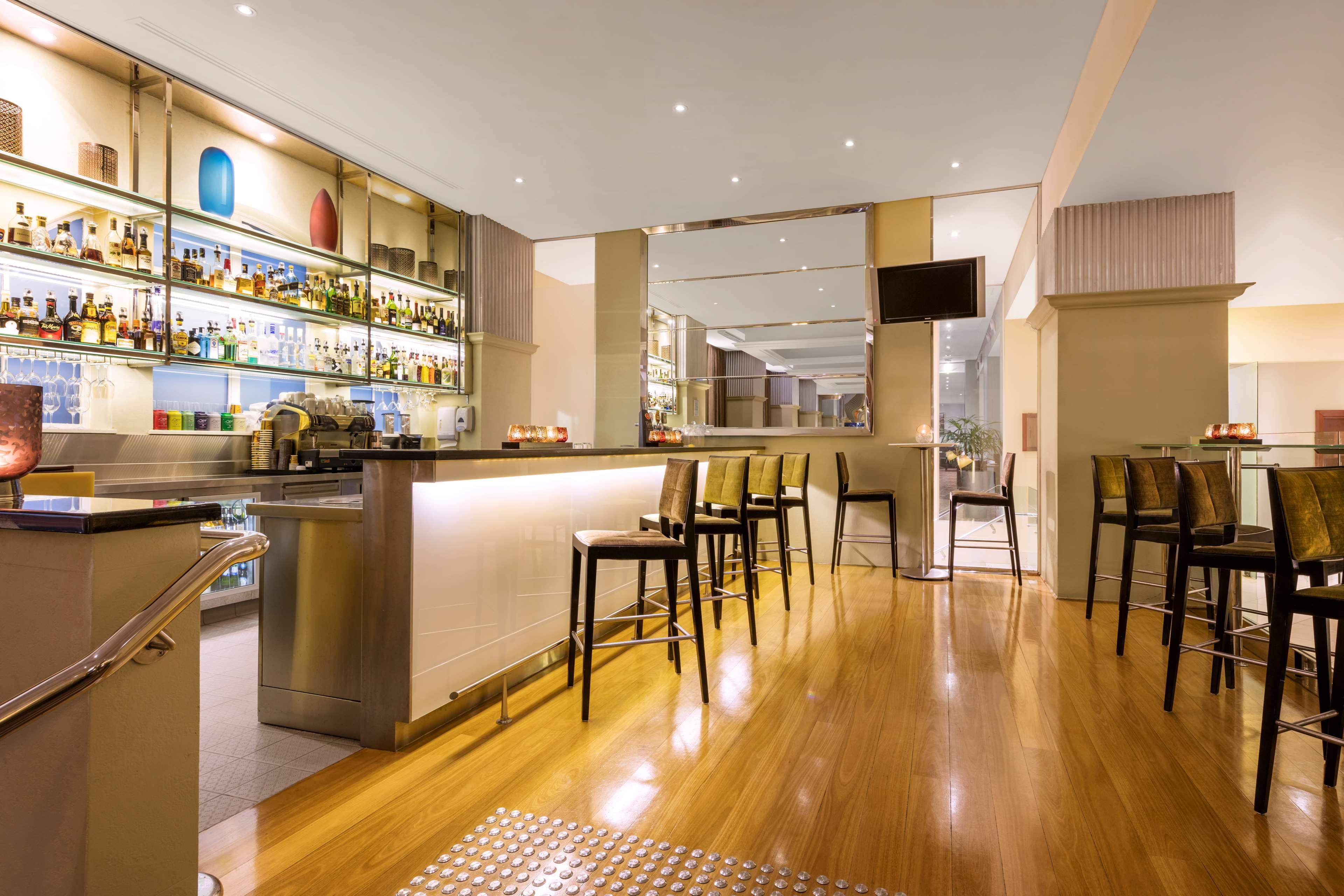 Radisson Blu Plaza Hotel ซิดนีย์ ภายนอก รูปภาพ The photo shows the interior of a modern bar or lounge area. There is a sleek bar counter with several high-backed stools arranged around it. Behind the bar, various bottles of liquor and decorative items are displayed on shelves, illuminated by soft