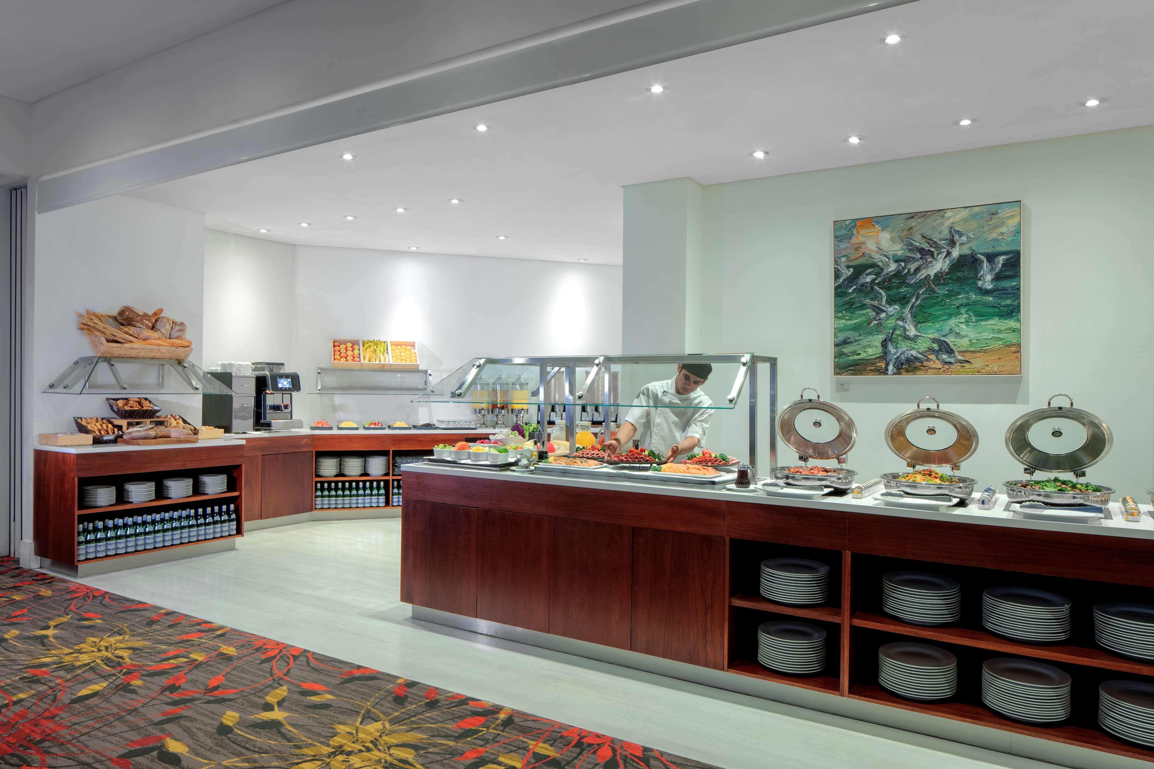 Radisson Blu Plaza Hotel ซิดนีย์ ภายนอก รูปภาพ The photo shows a buffet area in a dining space. There are several food stations arranged along a wall, featuring various dishes and offerings. A chef is visible behind the buffet, preparing food. The design includes wooden counters and shelves for s