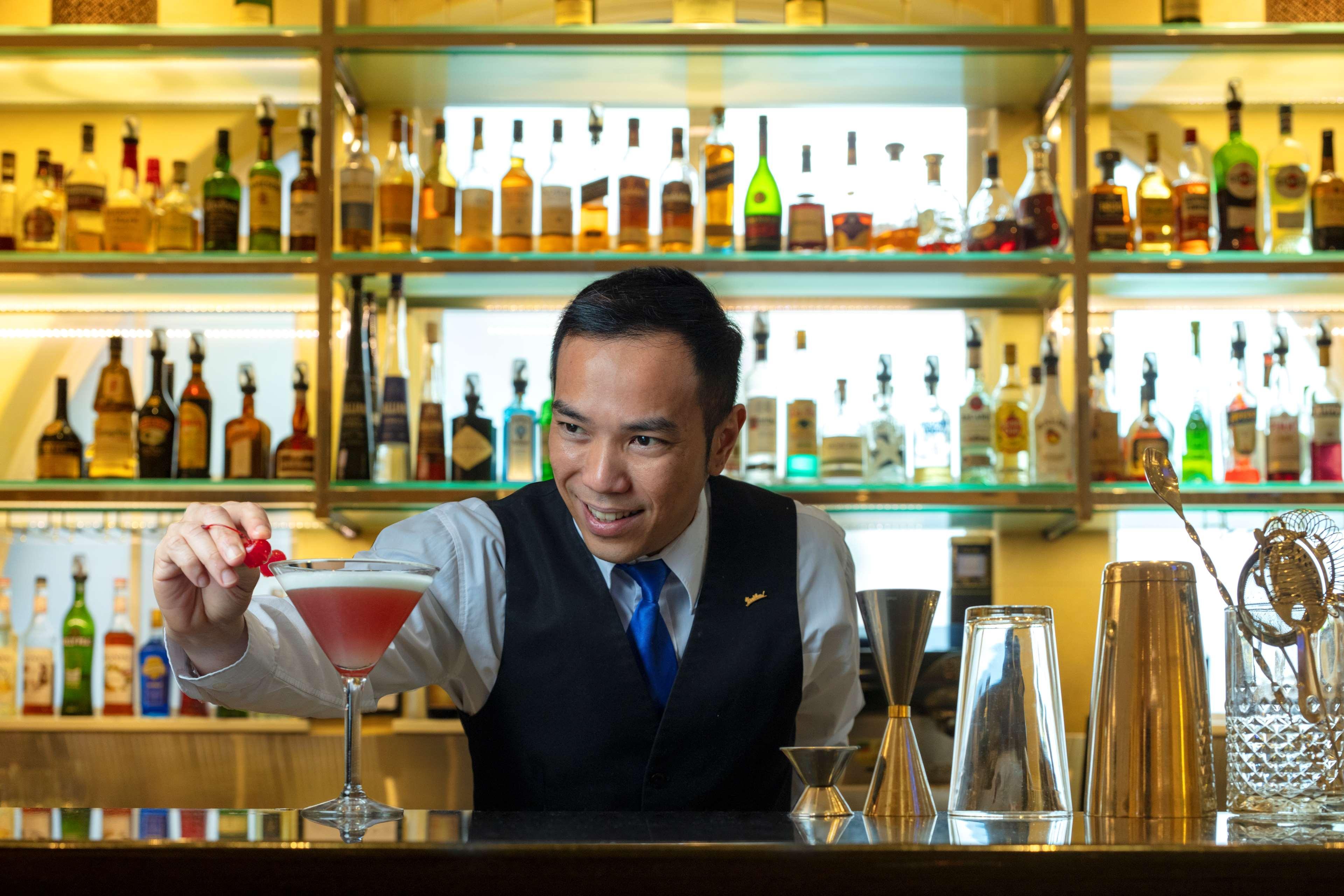 Radisson Blu Plaza Hotel ซิดนีย์ ภายนอก รูปภาพ The photo shows a bartender in a formal outfit, smiling as he prepares a cocktail. He is placing a garnish, possibly a cherry, on the rim of a elegantly presented glass filled with a pink drink. Behind him, there is a well-stocked bar with various bo