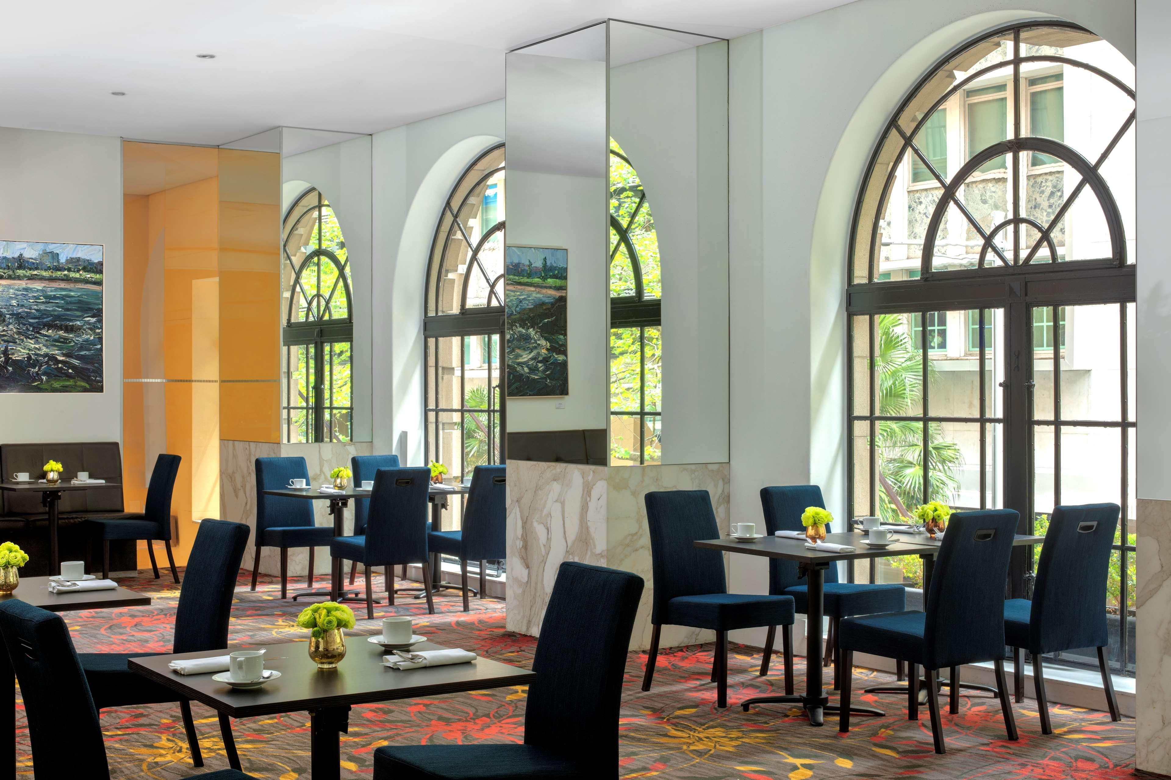 Radisson Blu Plaza Hotel ซิดนีย์ ภายนอก รูปภาพ The photo depicts a dining or lounge area with a modern and elegant design. Large arched windows allow natural light to fill the space, creating a bright and inviting atmosphere. The furnishings include dark, stylish chairs and tables, some configure