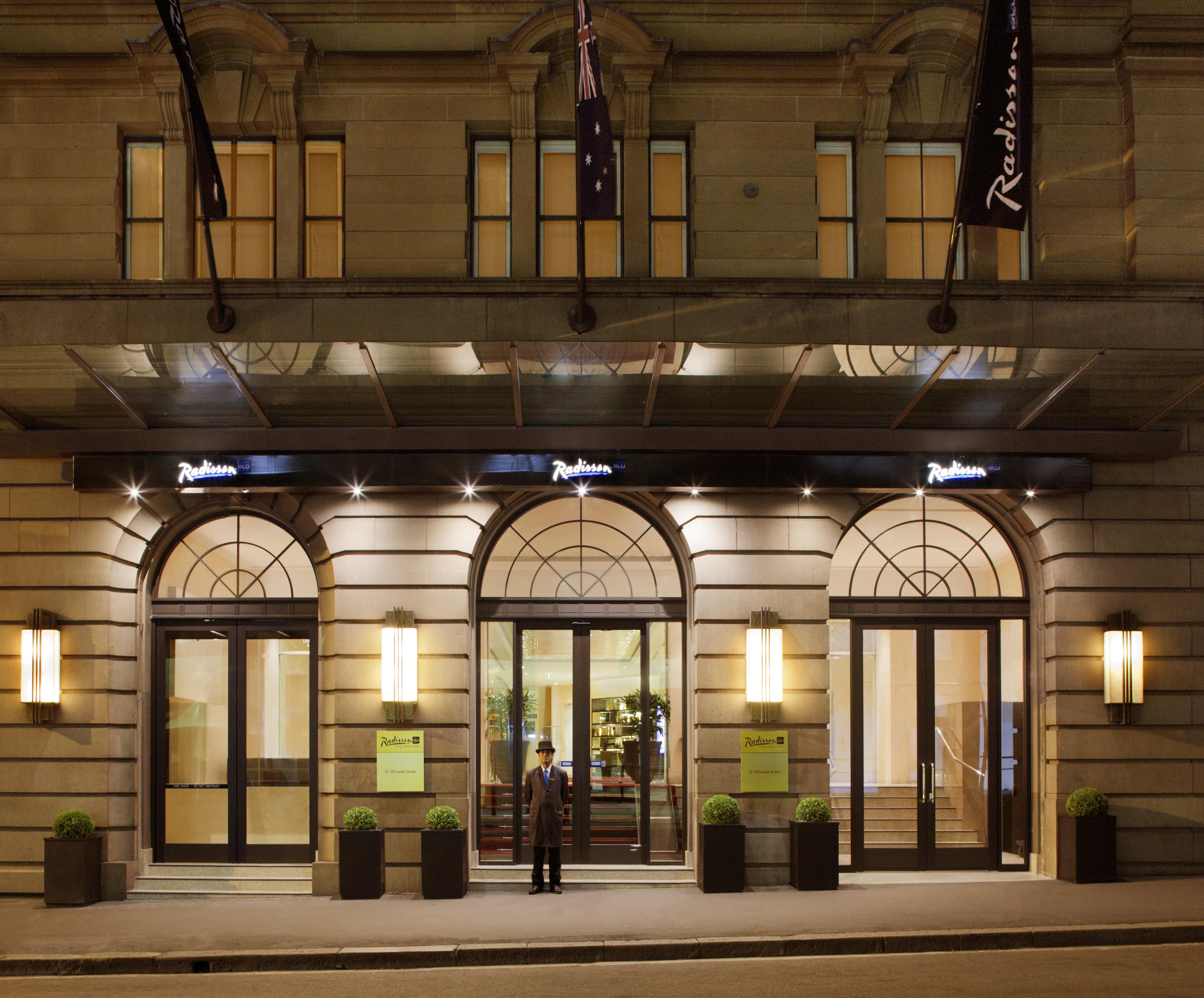 Radisson Blu Plaza Hotel ซิดนีย์ ภายนอก รูปภาพ The photo depicts the entrance of a Radisson hotel. The building features elegant architecture, with large arched windows and a well-lit canopy above the entrance. There are two potted plants on either side of the doors, which are framed by modern li