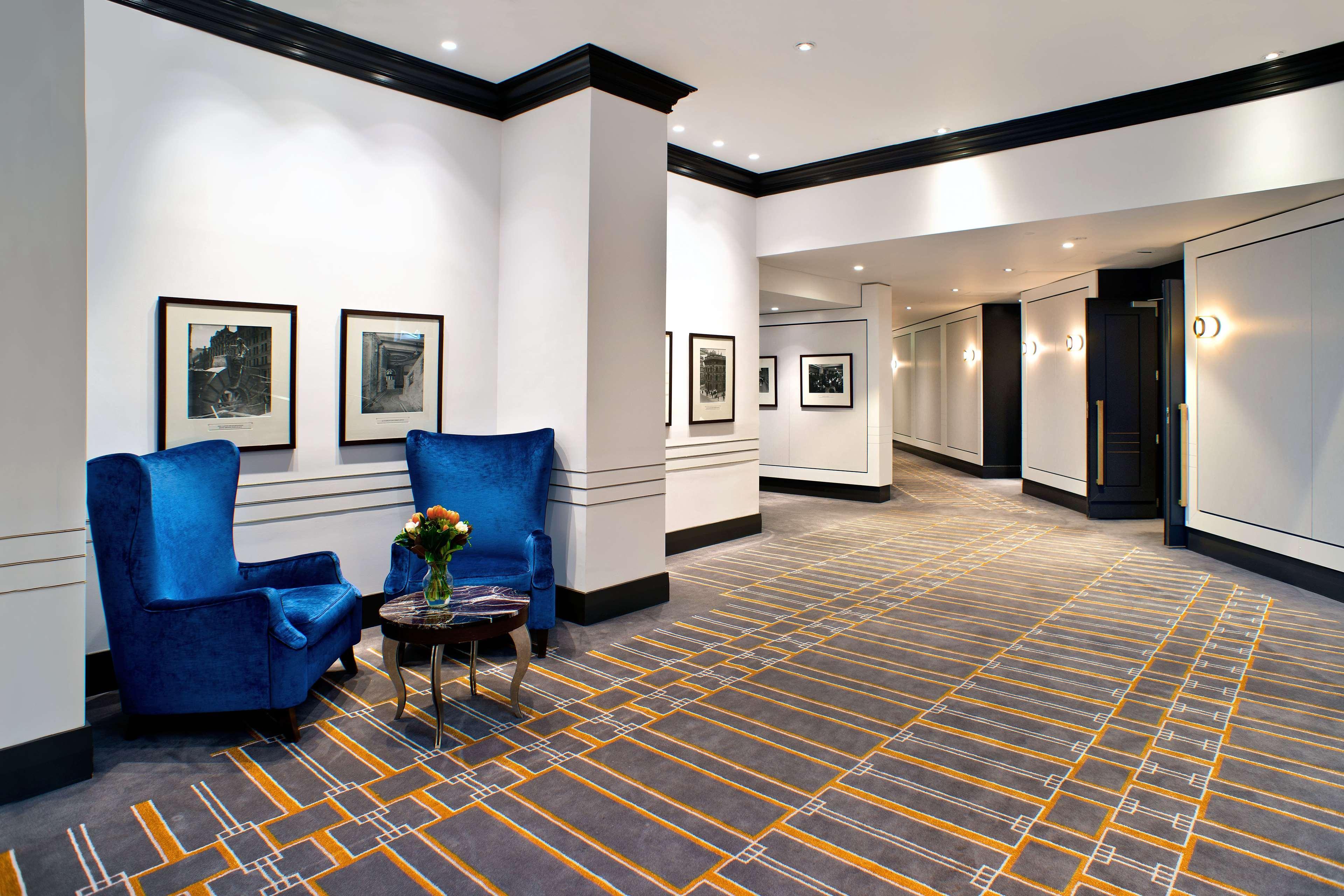 Radisson Blu Plaza Hotel ซิดนีย์ ภายนอก รูปภาพ The photo shows a modern hotel or building hallway. On one side, there are two vibrant blue armchairs arranged around a small table with a vase of flowers on it. The walls feature a few framed pictures, possibly black and white photographs. The floor