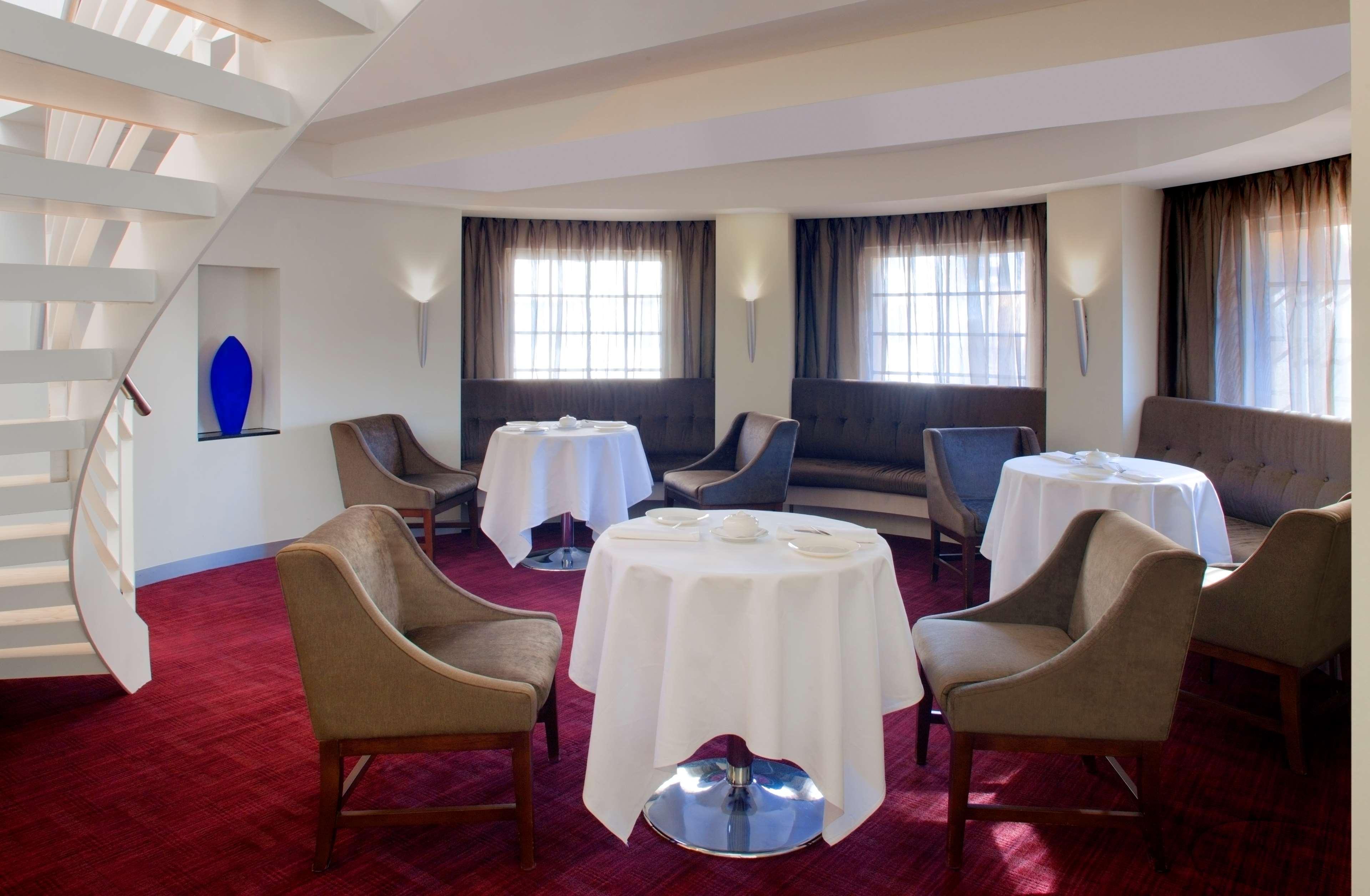 Radisson Blu Plaza Hotel ซิดนีย์ ร้านอาหาร รูปภาพ The photo shows a stylish dining area with a modern design. It features several round tables, each draped with white tablecloths. The seating consists of comfortable chairs with a neutral color, complemented by a plush bench along one wall. The space