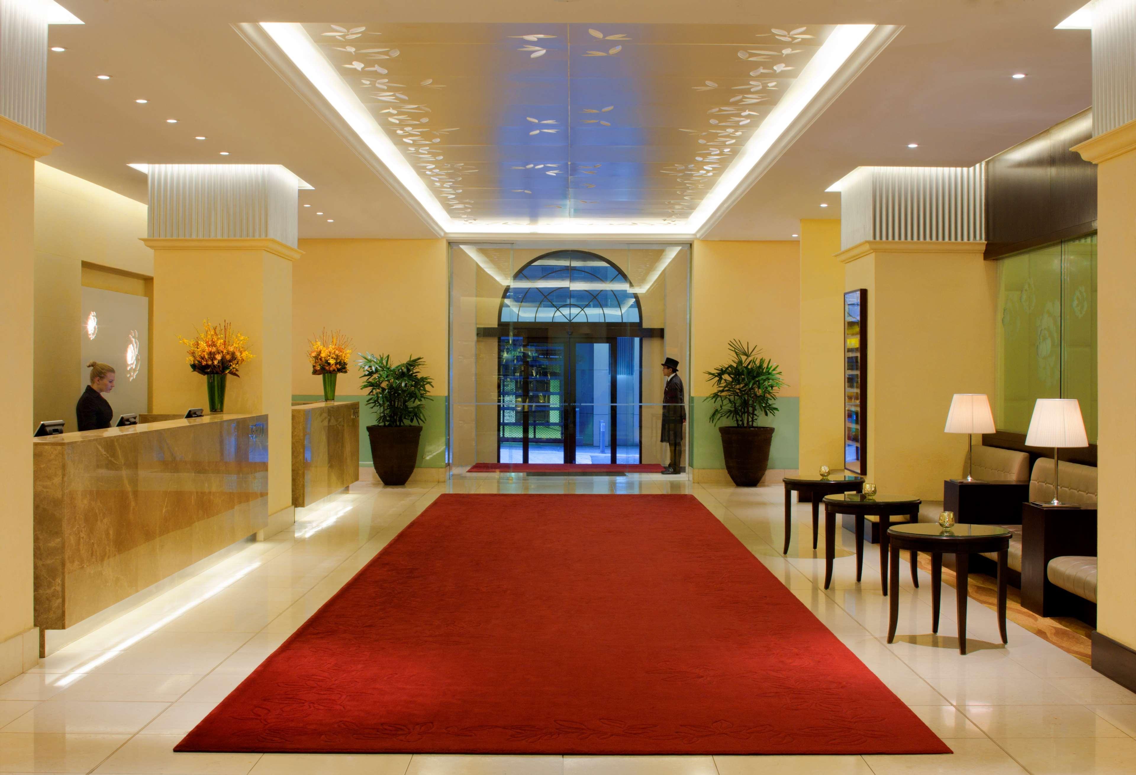 Radisson Blu Plaza Hotel ซิดนีย์ ภายใน รูปภาพ The photo shows the interior of a hotel lobby. It features a spacious and modern design with a warm color palette. There is a polished reception desk on the left, along with decorative plants. The floor is covered with a large red carpet that leads t
