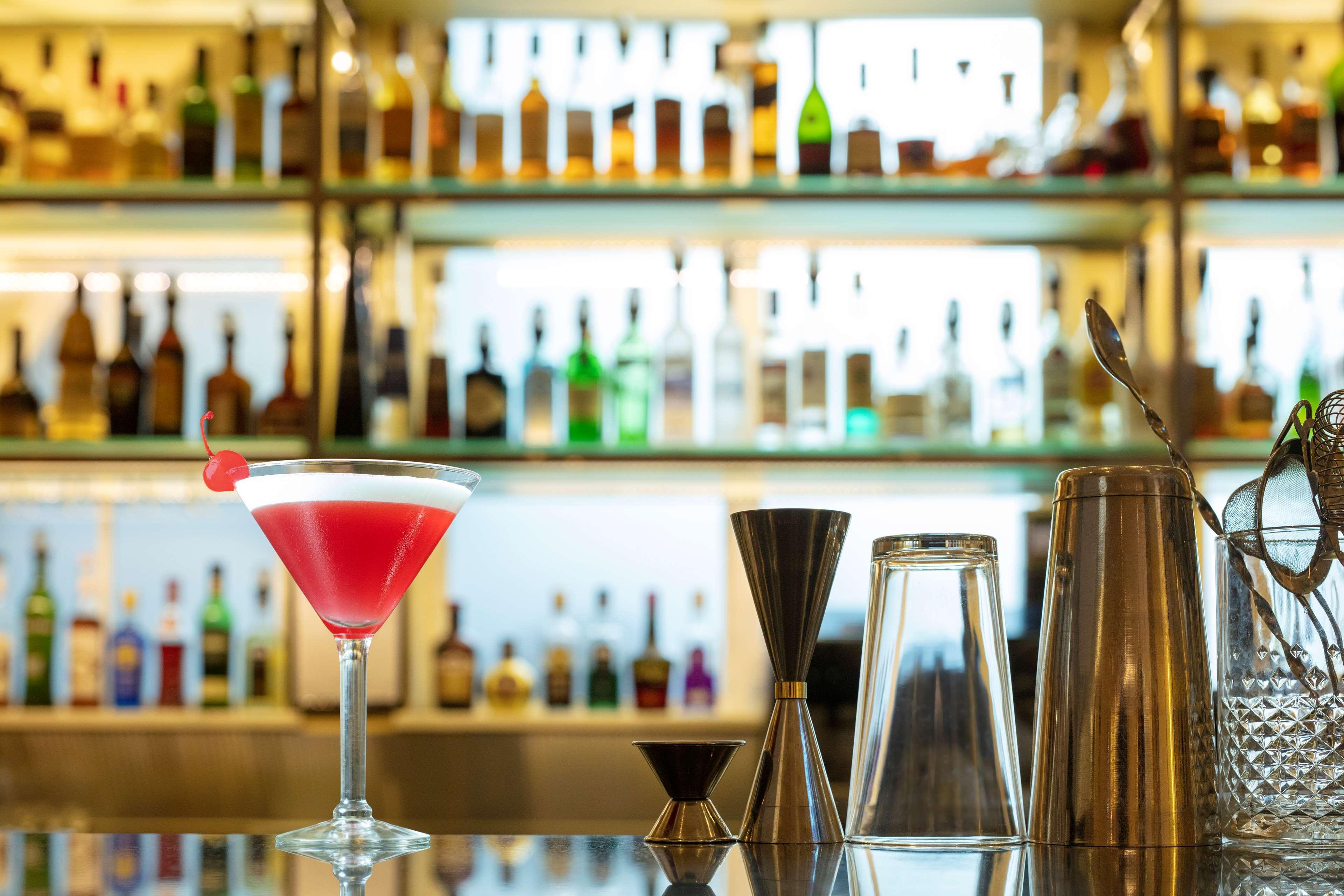 Radisson Blu Plaza Hotel ซิดนีย์ ภายนอก รูปภาพ The photo features a stylish bar setup. In the foreground, there is a tall cocktail glass filled with a pink drink, garnished with a cherry. Next to the glass, there are various bar tools, including a jigger for measuring, a shaker, and a clear glass