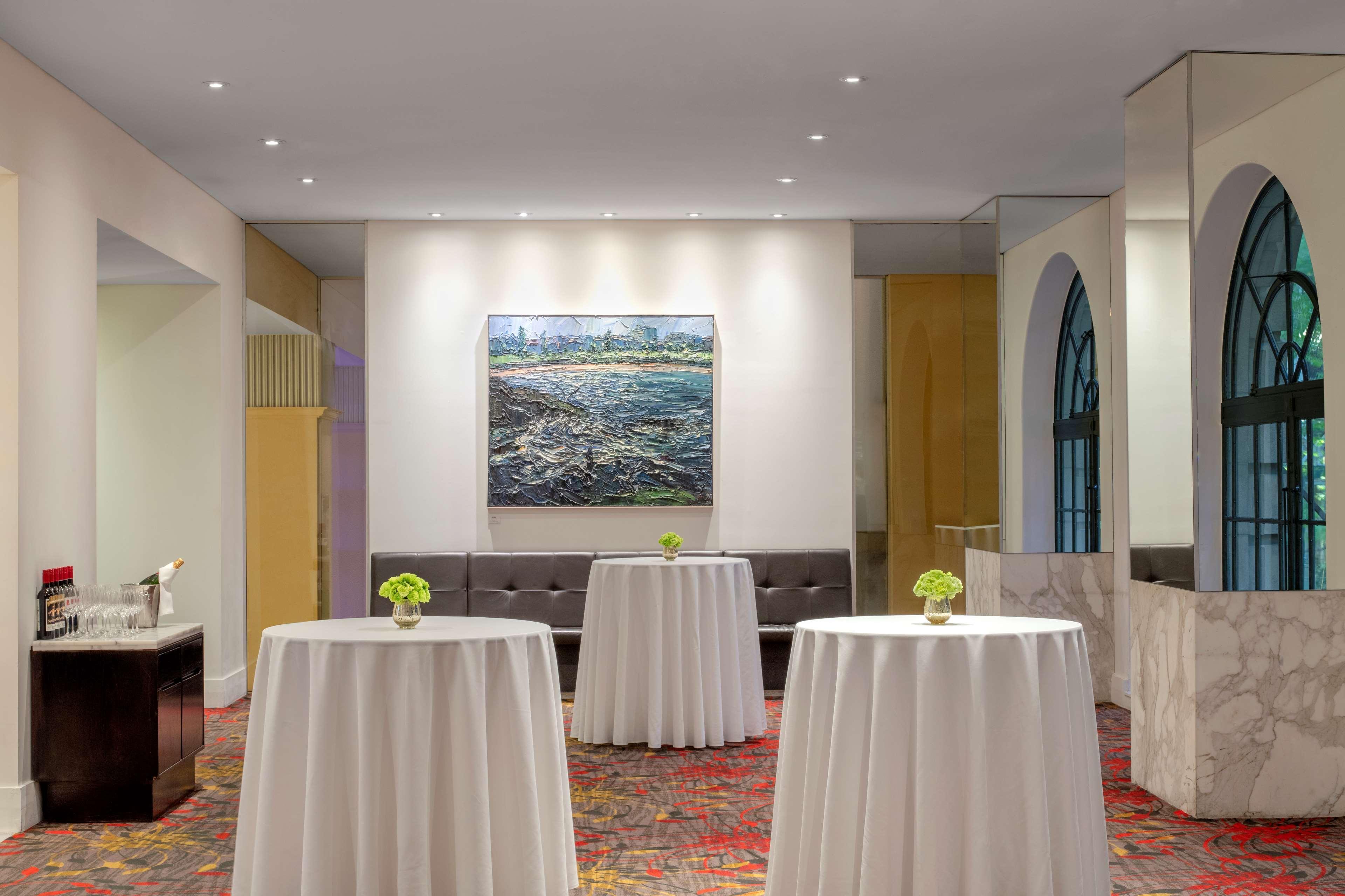 Radisson Blu Plaza Hotel ซิดนีย์ ภายนอก รูปภาพ The photo shows a modern event space. In the center, there are several round tables covered with white tablecloths, each topped with a small green floral arrangement. Behind the tables, there is a brown upholstered bench along the wall. A large paint
