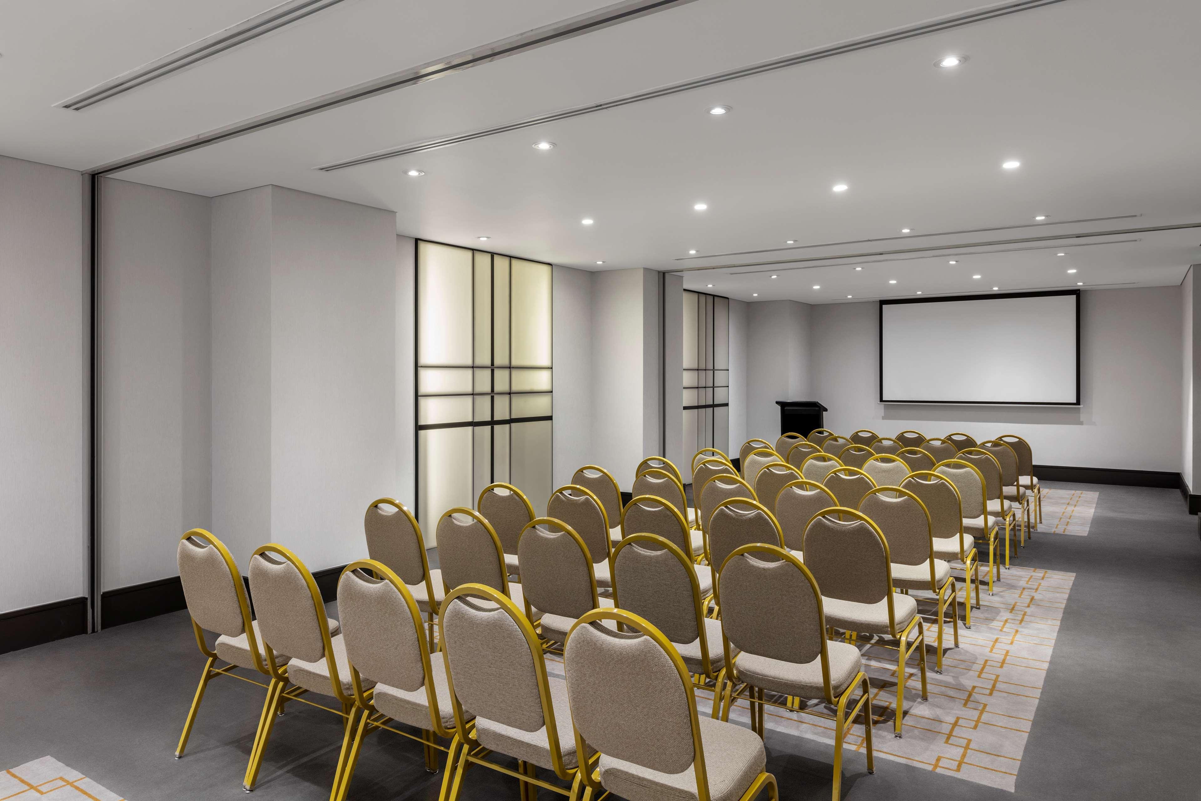 Radisson Blu Plaza Hotel ซิดนีย์ ภายนอก รูปภาพ The photo shows a conference room or meeting space. It features a neat arrangement of rows of beige chairs with gold frames facing a projection screen at the front. The room is well-lit with recessed lighting and has a modern design, including a clea