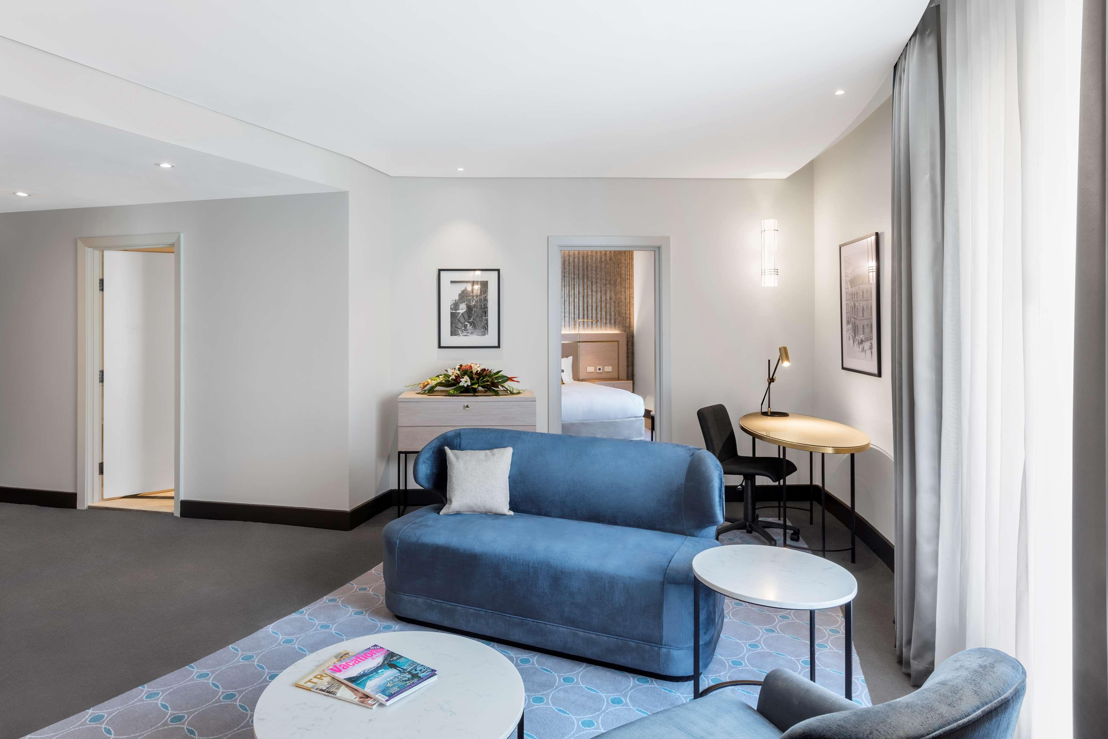 Radisson Blu Plaza Hotel ซิดนีย์ ภายนอก รูปภาพ The photo depicts a modern hotel room or suite that features a comfortable and stylish living area. The room includes a light blue sofa complemented by a small round coffee table. There is a decorative pillow on the sofa. To the side, there is a circ