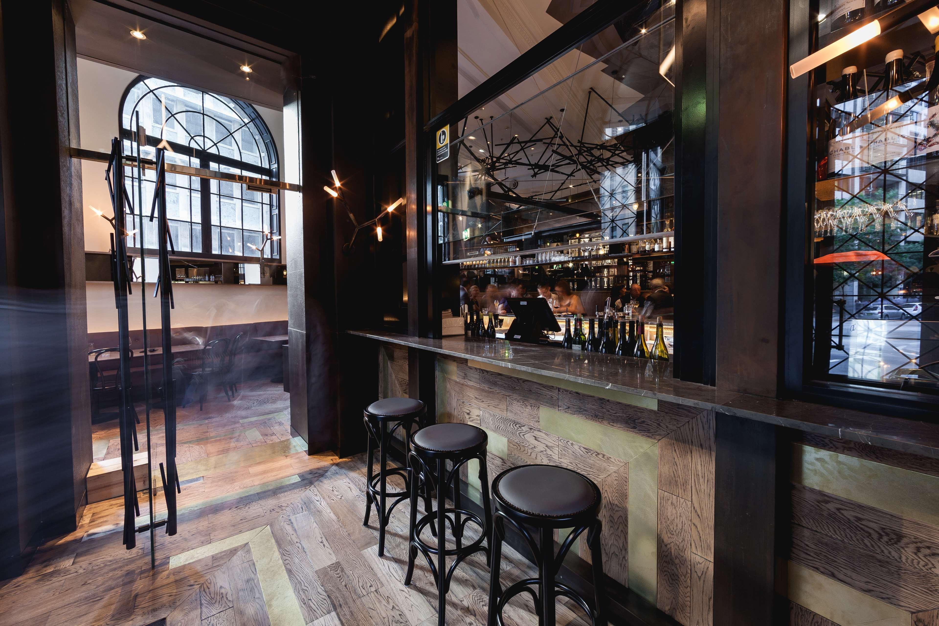 Radisson Blu Plaza Hotel ซิดนีย์ ภายนอก รูปภาพ The photo shows the interior of a stylish bar or lounge. There are several tall, black stools arranged in front of a polished bar counter. The bar has a glass backing that showcases a variety of bottles, indicating a well-stocked bar area. The floori