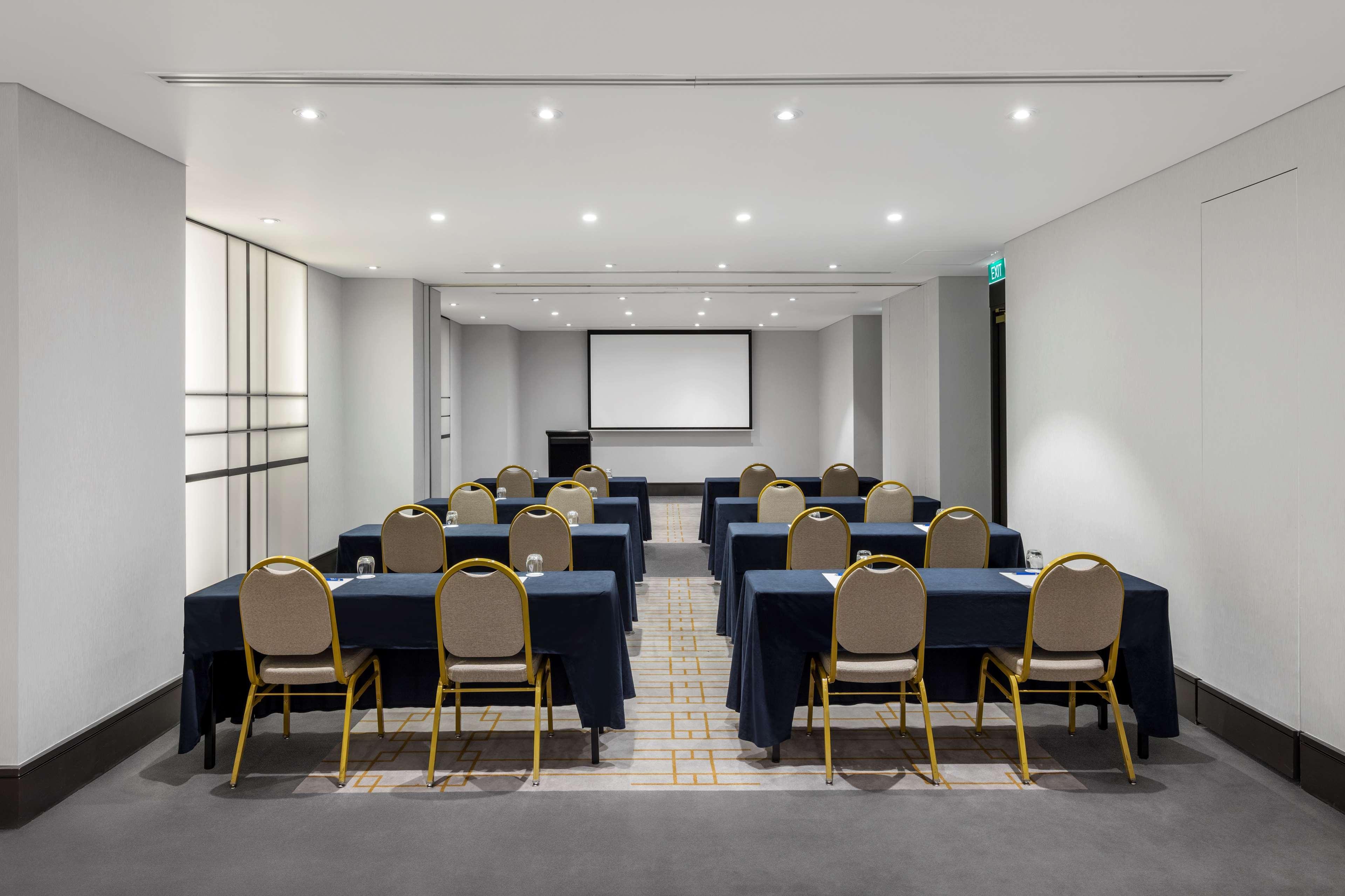 Radisson Blu Plaza Hotel ซิดนีย์ ภายนอก รูปภาพ The photo shows a conference room set up for a meeting or presentation. There are several tables arranged in a U-shape, each covered with dark blue tablecloths. Each table has a gold-colored chair positioned at it. At the front of the room, there is 