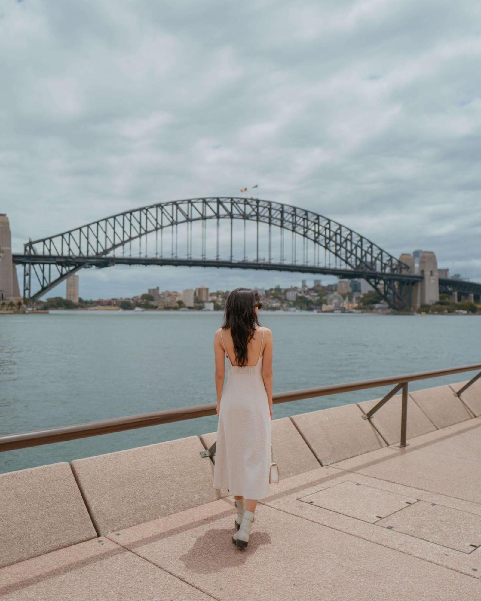 Radisson Blu Plaza Hotel ซิดนีย์ ภายนอก รูปภาพ The photo depicts a woman standing by a body of water, facing away from the camera. She is wearing a light-colored dress and appears to be looking at the Sydney Harbour Bridge, which is arched and spans across the water. The sky is overcast, suggesti