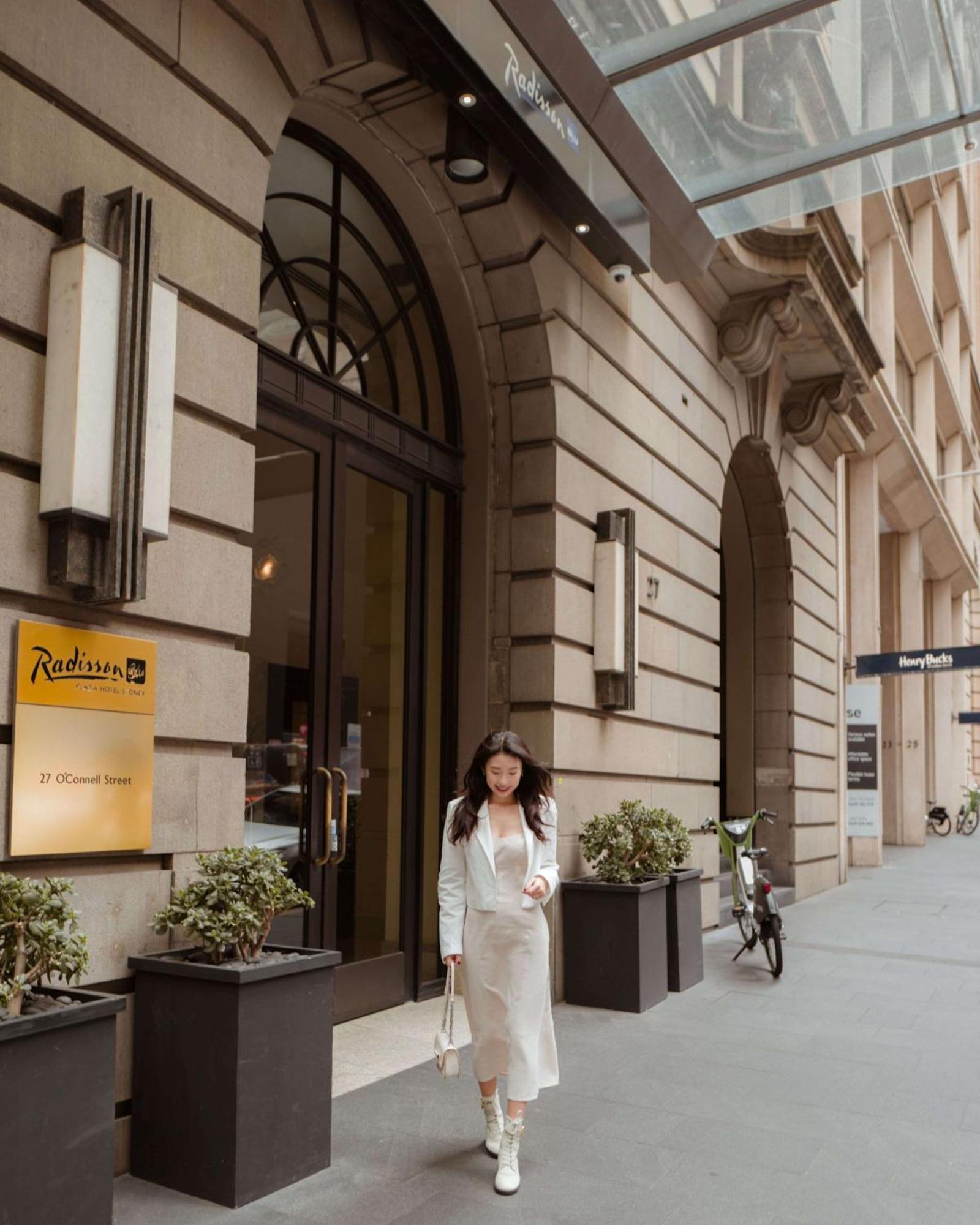 Radisson Blu Plaza Hotel ซิดนีย์ ภายนอก รูปภาพ The photo shows the exterior of a building, likely a hotel, with a sign indicating it is the Radisson. A woman dressed in a light-colored outfit is walking out of the entrance. There are potted plants on either side of the door, and in the background
