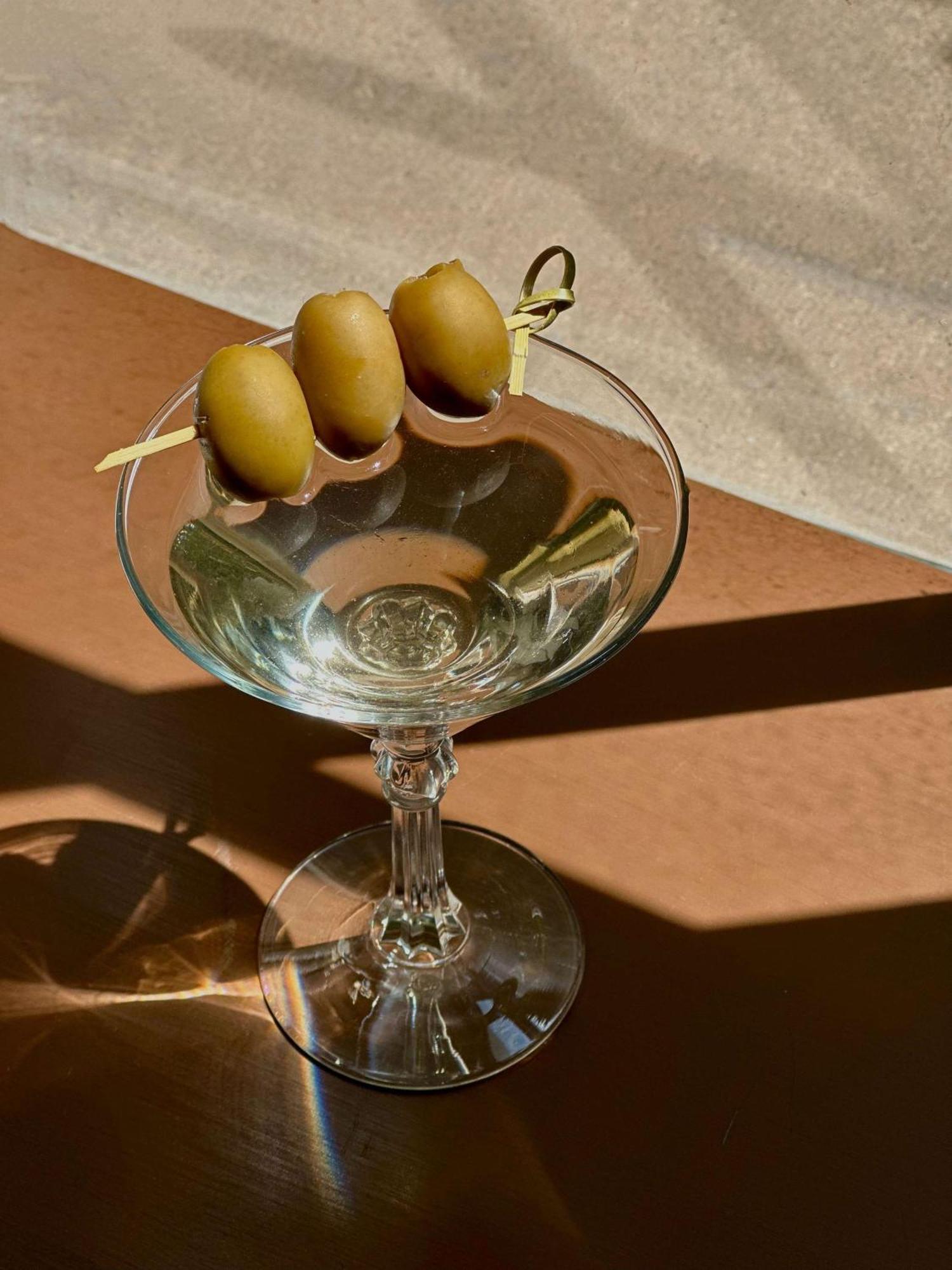 Radisson Blu Plaza Hotel ซิดนีย์ ภายนอก รูปภาพ The image shows a clear martini glass with three green olives skewered on a toothpick resting on the rim. The glass is empty, reflecting light and showcasing its elegant shape. The background features a warm-toned surface and possibly a shadow, enhan