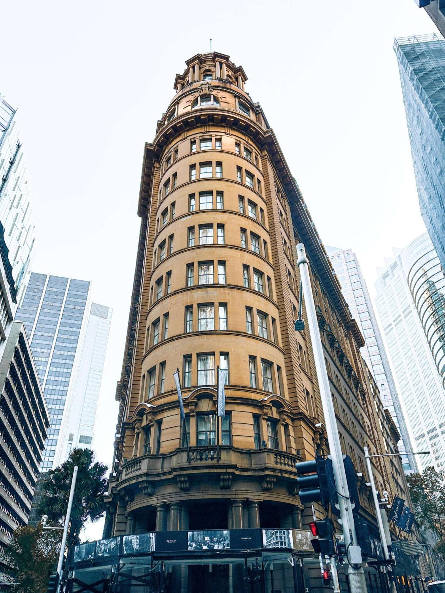 Radisson Blu Plaza Hotel ซิดนีย์ ภายนอก รูปภาพ The image shows a distinctive architectural building characterized by its curved facade and a warm, yellowish-brown exterior. The structure appears to have multiple floors, with large windows arranged symmetrically. Surrounding the building are moder