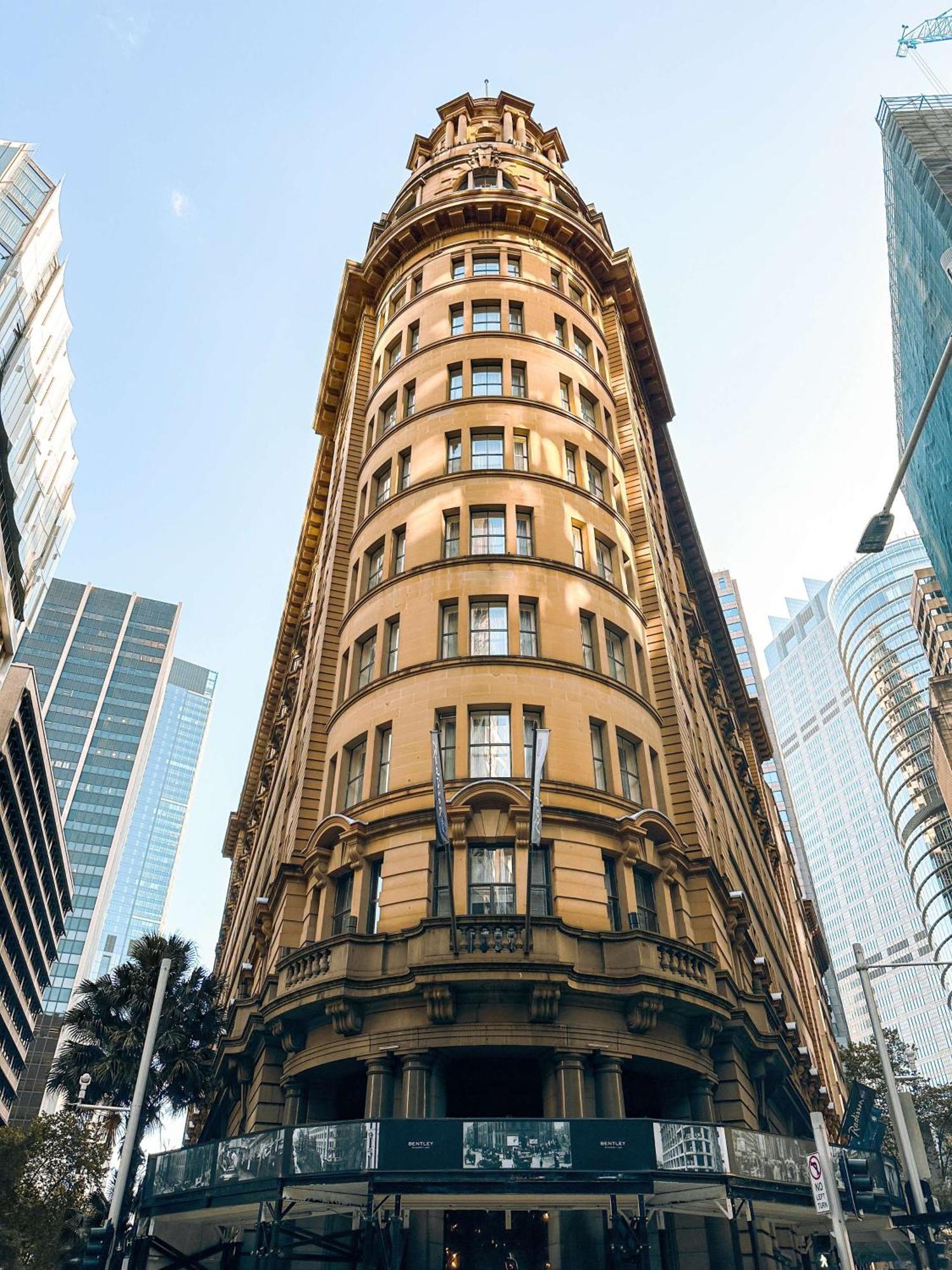 Radisson Blu Plaza Hotel ซิดนีย์ ภายนอก รูปภาพ The photo features a tall, historic building characterized by its rounded, cylindrical shape and numerous windows. The facade is ornate with a warm, yellowish-brown color, and the architecture reflects styles from an earlier era. Flanking the buildin