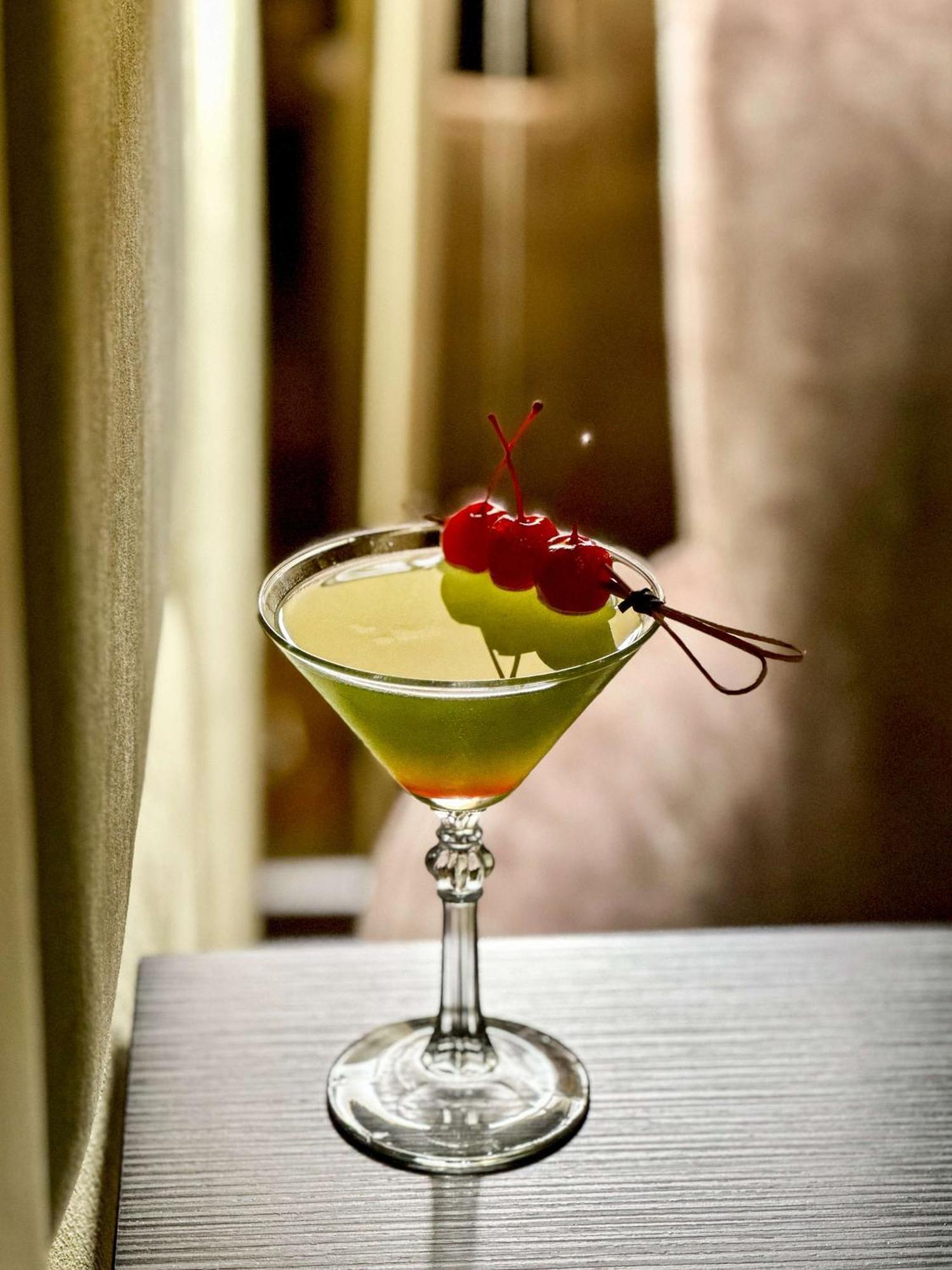 Radisson Blu Plaza Hotel ซิดนีย์ ภายนอก รูปภาพ The image shows a cocktail in a classic martini glass. The drink has a vibrant green color and is garnished with two yellow-green fruits (possibly a melon or similar) and three cherries on a skewer. The background appears blurred, creating a soft foc