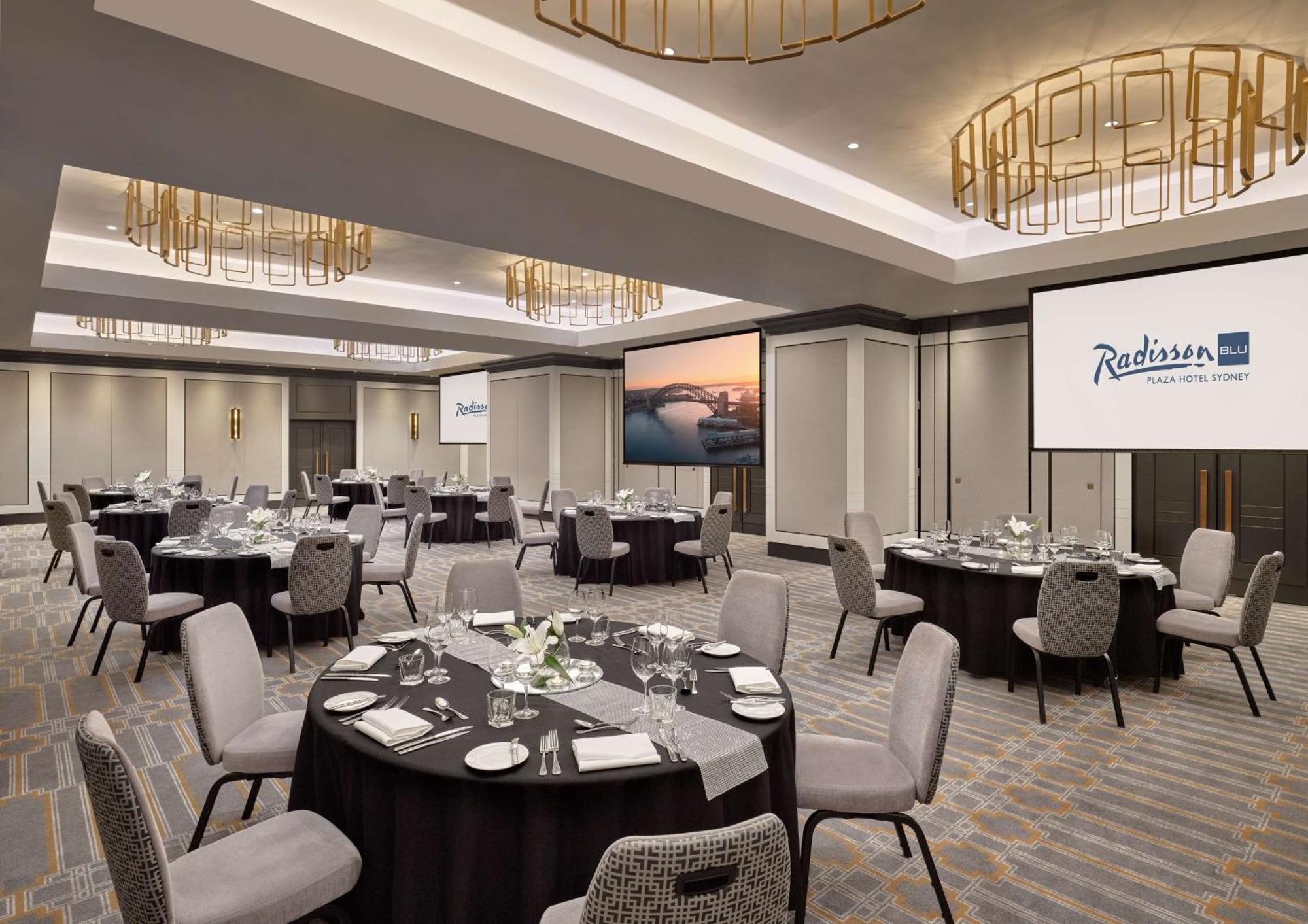 Radisson Blu Plaza Hotel ซิดนีย์ ภายนอก รูปภาพ The photo shows an elegant banquet or conference room setup at a Radisson hotel. The space features multiple round tables, each adorned with white tablecloths, centerpieces, and plates, ready for dining. The room is well-lit, highlighting modern deco