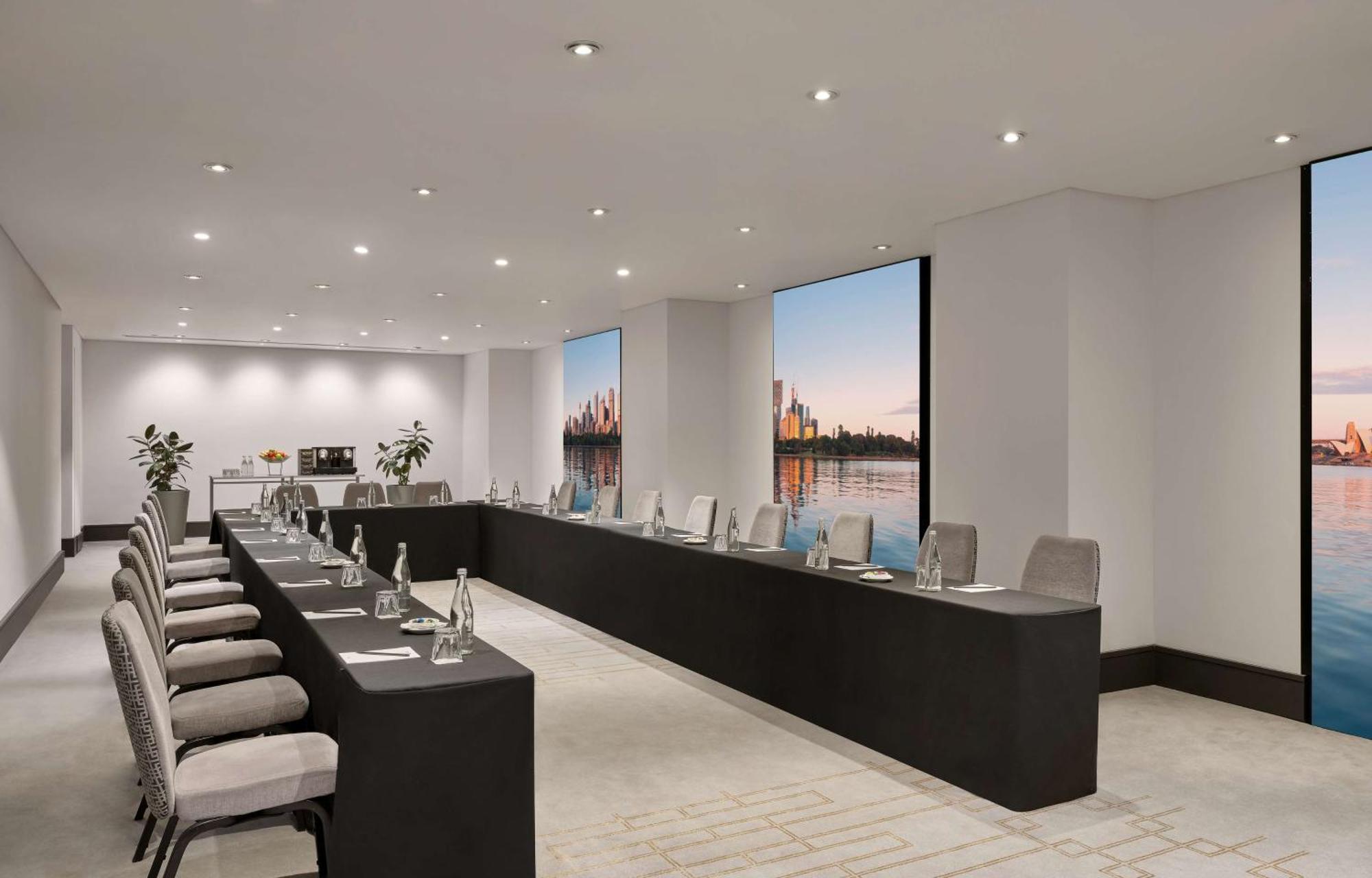 Radisson Blu Plaza Hotel ซิดนีย์ ภายนอก รูปภาพ The photo depicts a modern conference room set up for a meeting. The room features a long, rectangular table with multiple chairs arranged along its sides. Each chair is equipped with items such as water bottles and notepads. Large windows allow natu