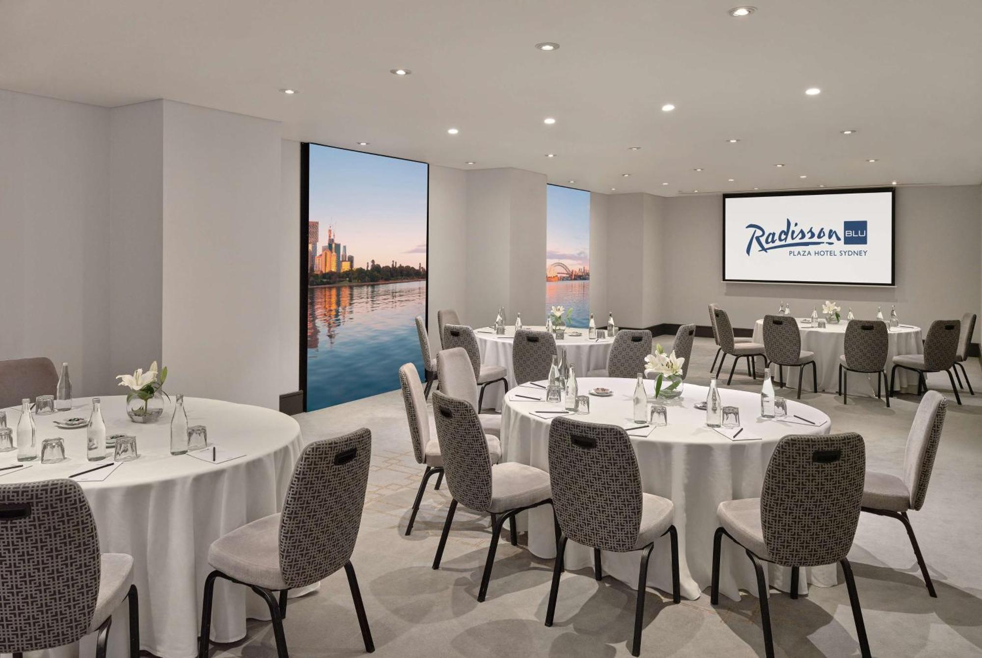Radisson Blu Plaza Hotel ซิดนีย์ ภายนอก รูปภาพ The photo shows a modern meeting room at the Radisson Hotel in a city. The room features several round tables set with white tablecloths and gray upholstered chairs. There are small flower arrangements on each table, creating a welcoming atmosphere. 
