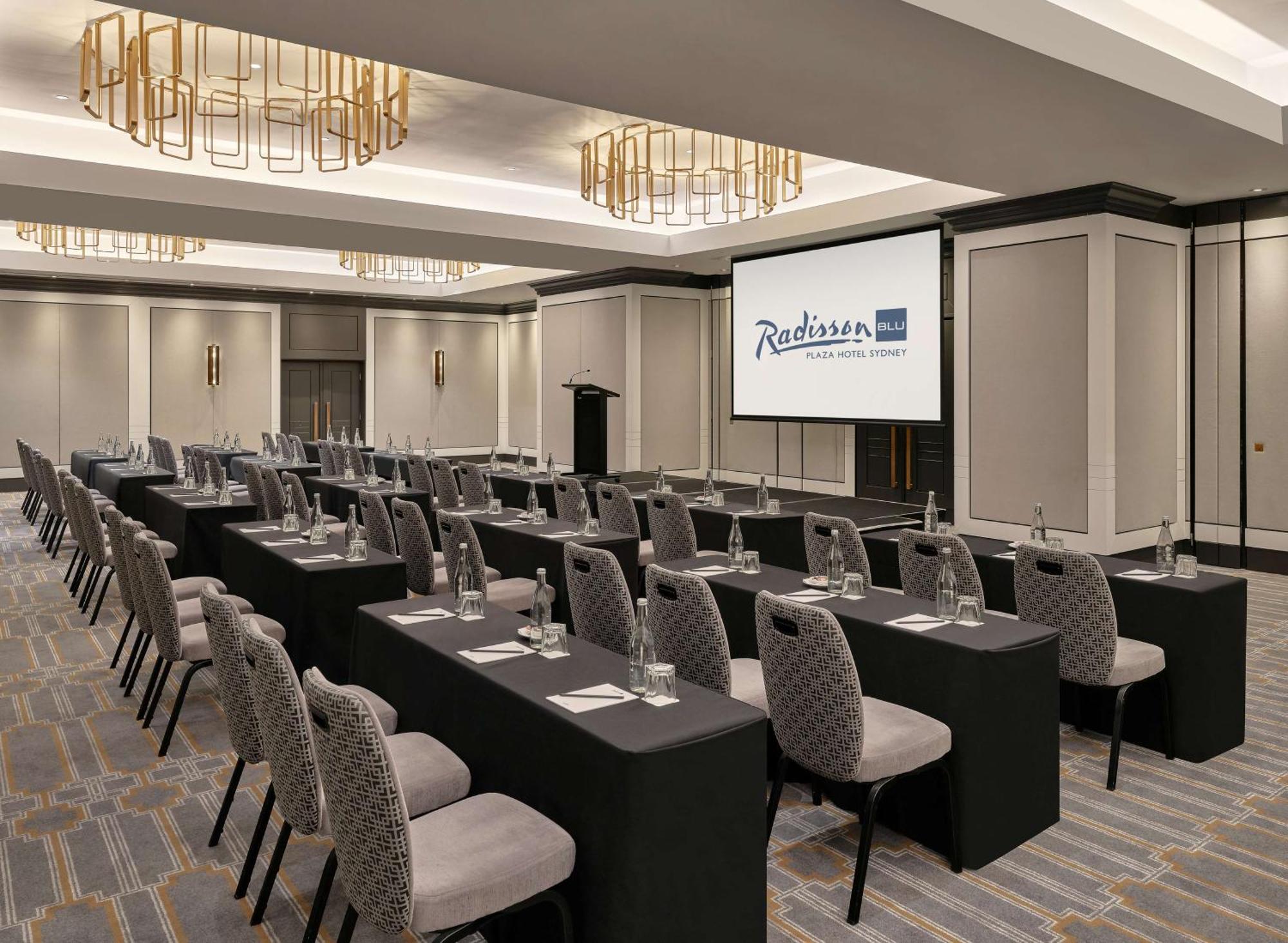 Radisson Blu Plaza Hotel ซิดนีย์ ภายนอก รูปภาพ The photo shows a conference or meeting room set up in a hotel, likely a Radisson. The room features several rows of tables covered in black tablecloths, each with a chair beside it. Water glasses are placed on each table, and there appears to be a l